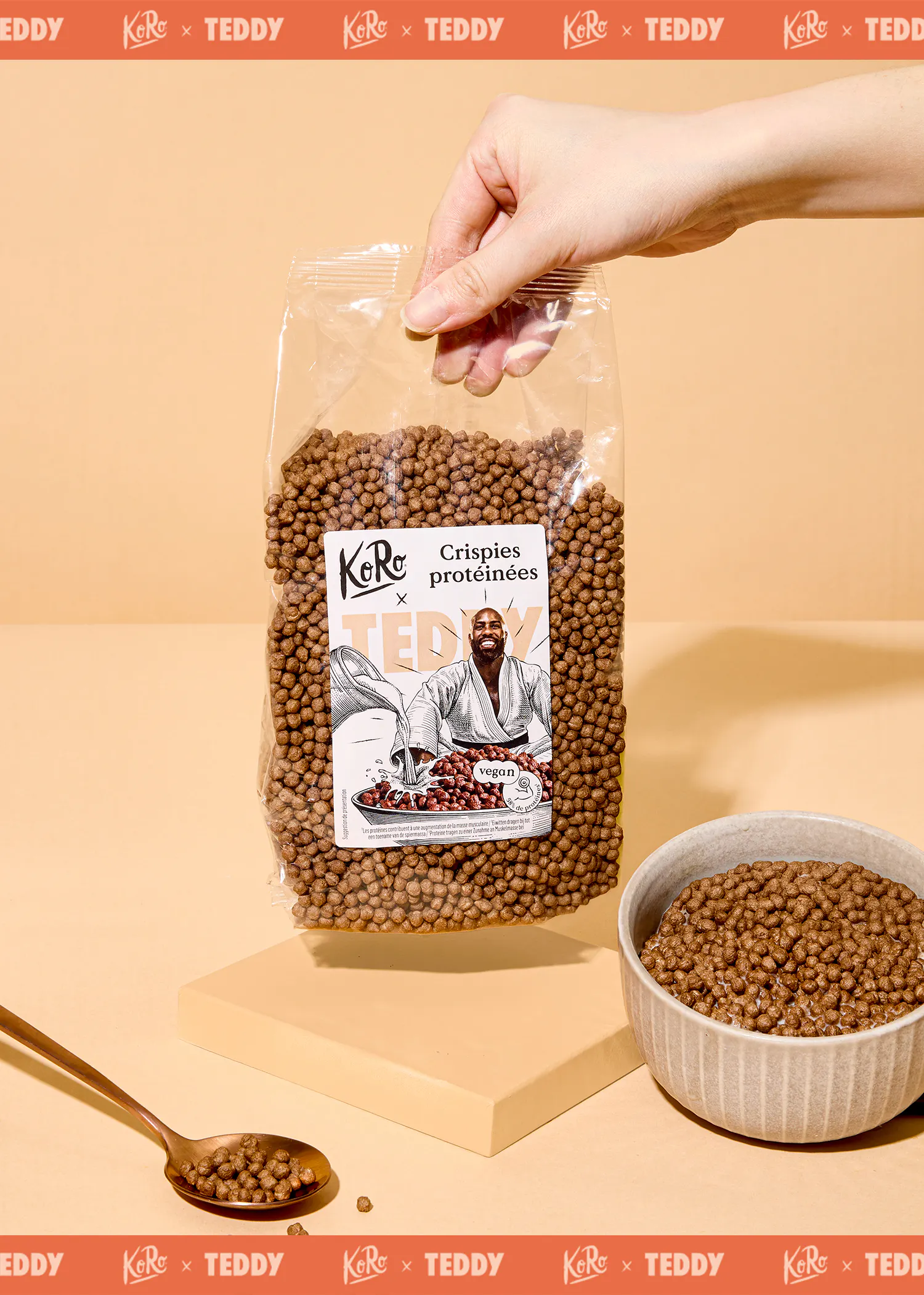 une main tient un sac de crispies protéinées koro x teddy avec l'illustration d'un homme en judogi et la mention "vegan", posé à côté d'un bol rempli de crispies et d'une cuillère contenant des crispies.