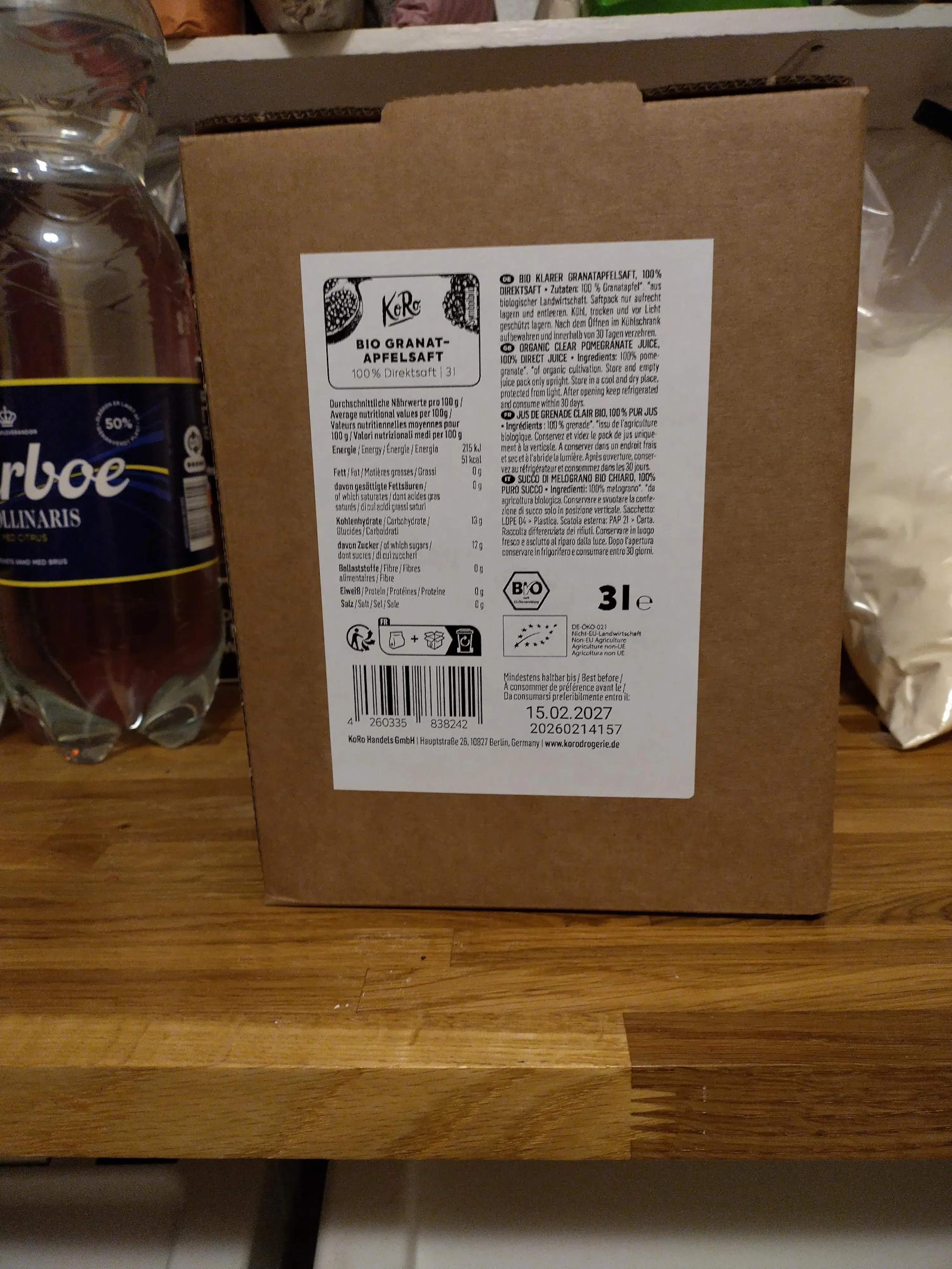 eine braune kartonverpackung von koro bio granatapfelsaft (100% direktsaft, 3l) mit nährwertangaben und dem haltbarkeitsdatum 15.02.2027 ist auf einem holztisch zu sehen.