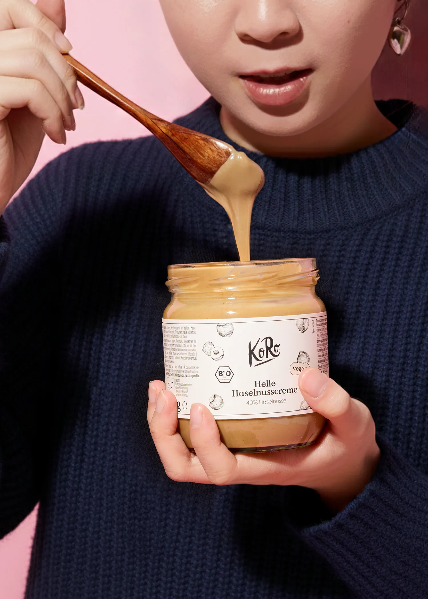 eine person hält ein glas koro helle haselnusscreme, aus dem ein löffel mit haselnusscreme tropft.