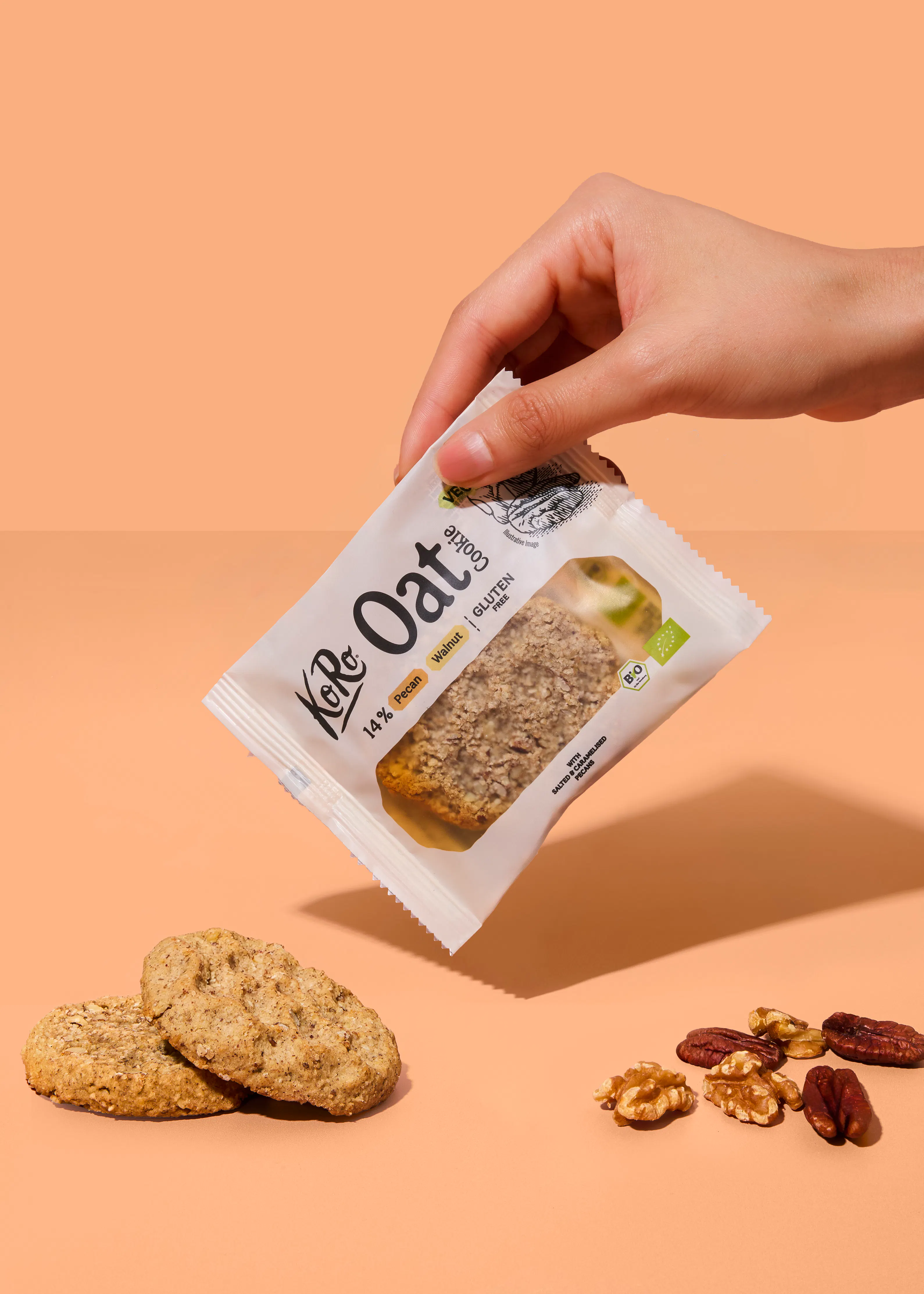 a hand holds a koro oat cookie package above two whole cookies and scattered walnut and pecan pieces on a light brown background.