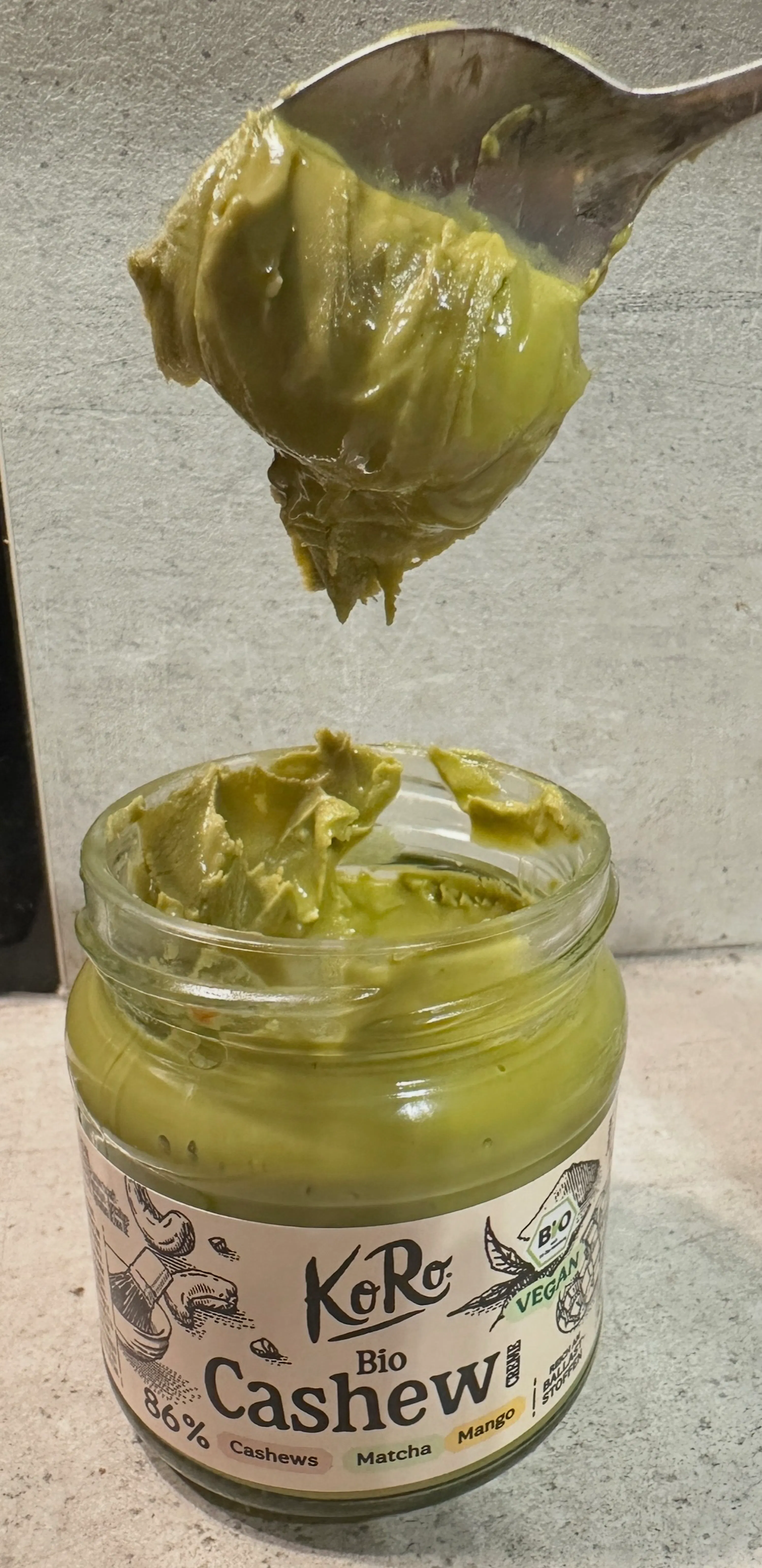 ein löffel hebt grüne cashew-matcha-mango-butter aus einem glas mit dem etikett "koro bio cashew matcha mango".