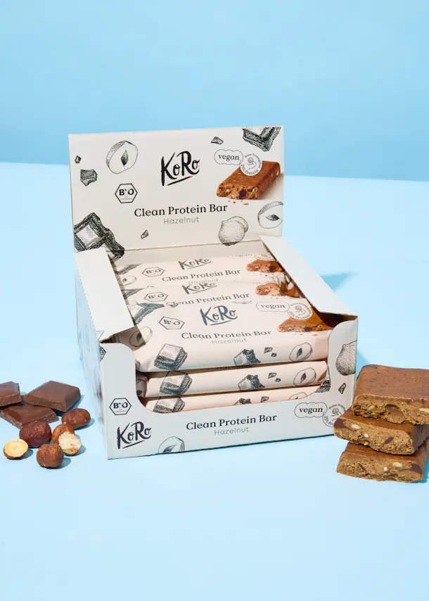 eine offene displaybox mit mehreren verpackten koro clean protein riegeln haselnuss, die als vegan gekennzeichnet sind, daneben liegen lose haselnüsse, schokoladenstücke und ein angeschnittener riegel.