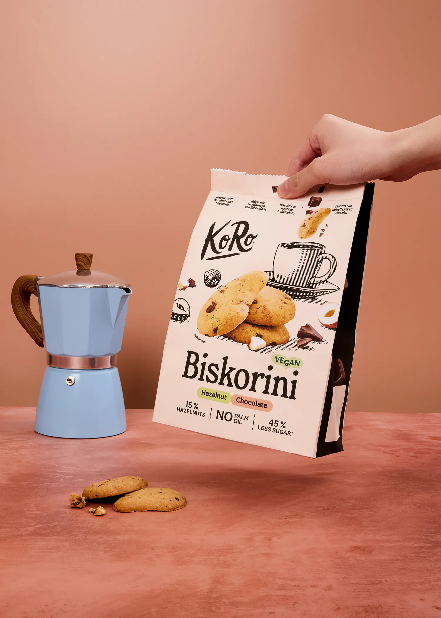 una mano sostiene un paquete de galletas biskorini veganas de avellana y chocolate, junto a una cafetera moka azul y galletas sueltas sobre una superficie rosa.