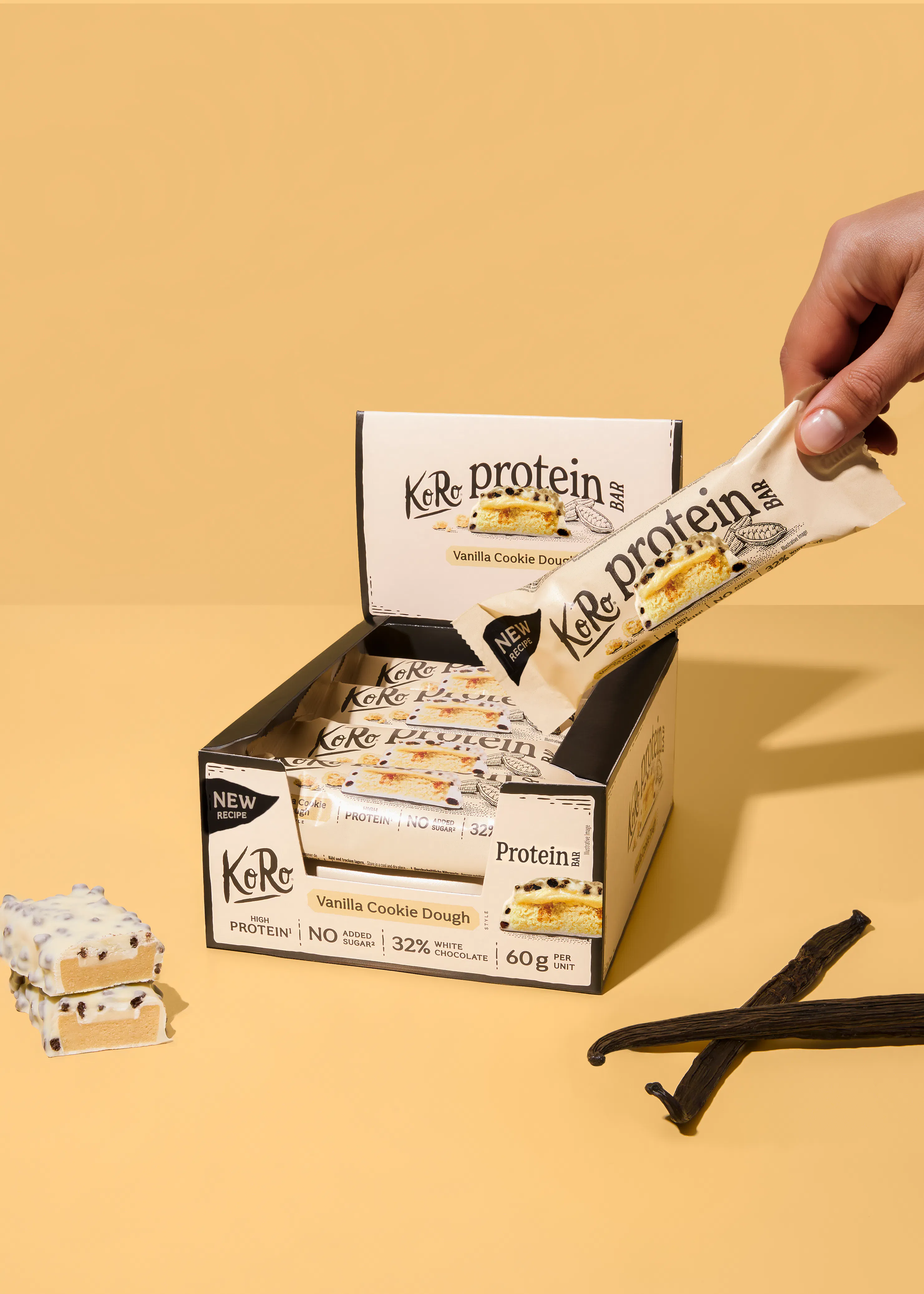 a hand reaches into a display box to grab a koro protein vanilla cookie dough bar, with two unwrapped bars and vanilla beans visible on a yellow background.