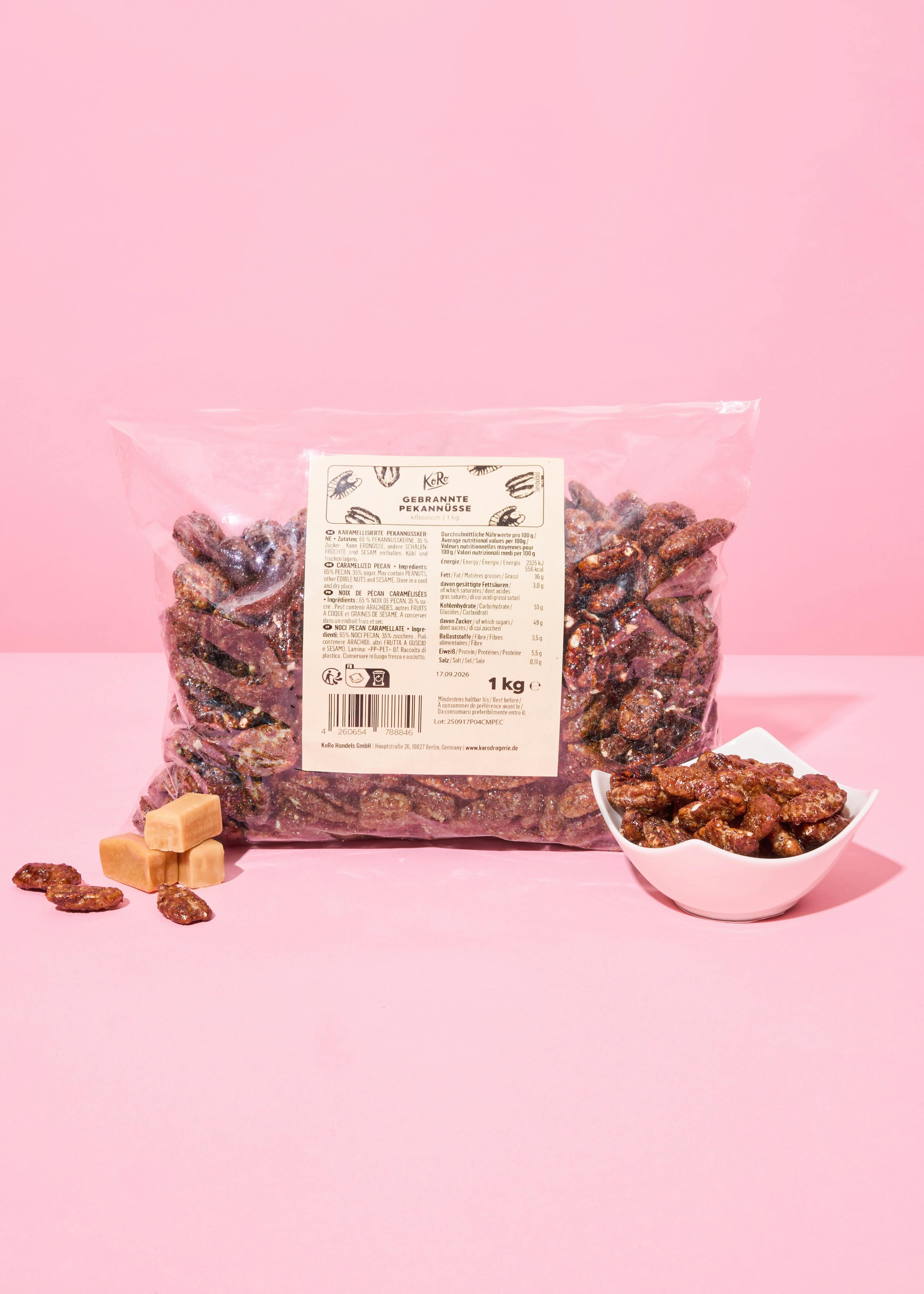 un grand sac transparent de noix de pécan caramélisées, avec un petit bol de noix de pécan et trois bonbons au caramel à côté, sur un fond rose.