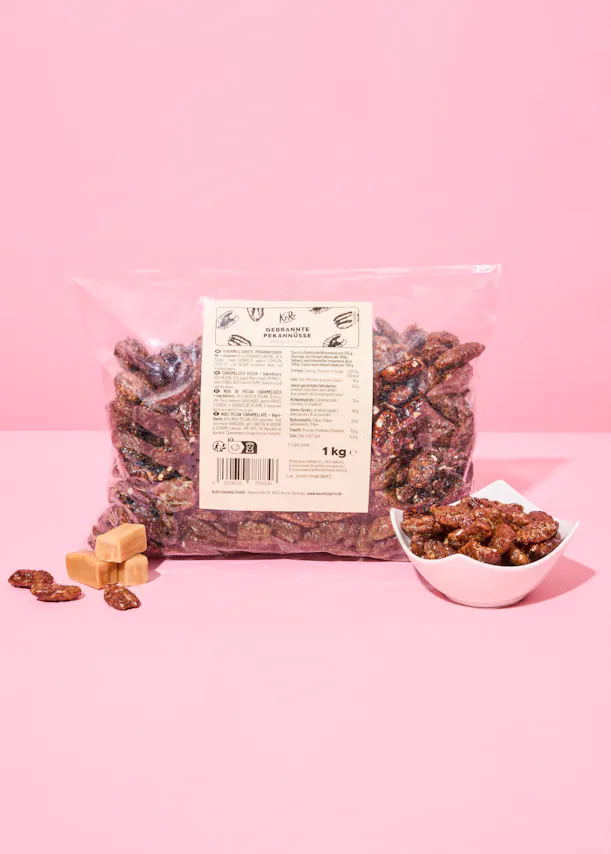 un grand sac transparent de noix de pécan caramélisées, avec un petit bol de noix de pécan et trois bonbons au caramel à côté, sur un fond rose.