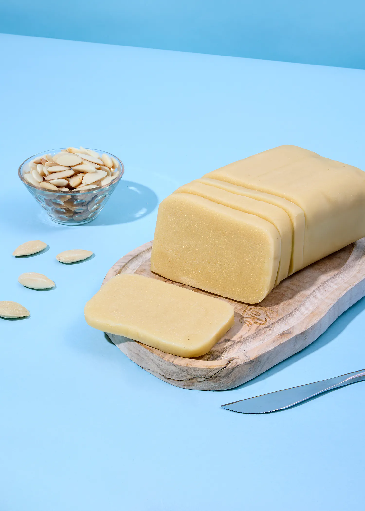 ein teilweise geschnittener marzipanblock auf einem holzbrett, daneben eine schale mit mandelsplittern und ein messer auf blauem hintergrund.