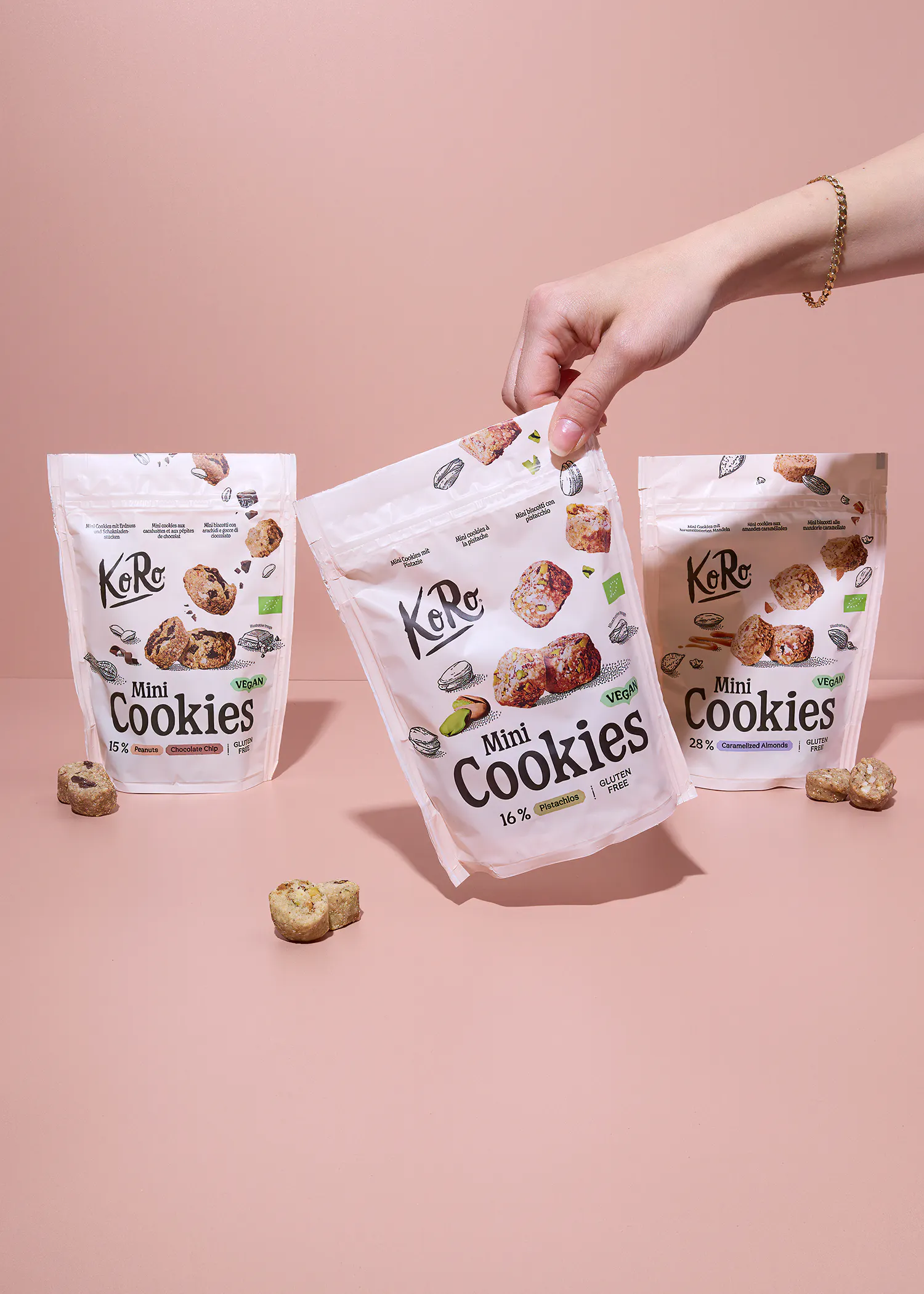 eine hand hält einen beutel koro mini cookies mit pistazien, während zwei weitere beutel mit erdnuss-schokochip und karamellisierten mandeln sowie einzelne kekse auf einem rosa hintergrund zu sehen sind.
