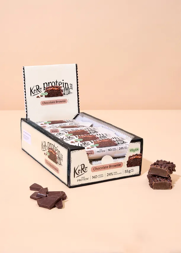 an open display box of koro protein chocolate brownie bars, with loose chocolate pieces and a cut bar showing its filling, on a peach background.