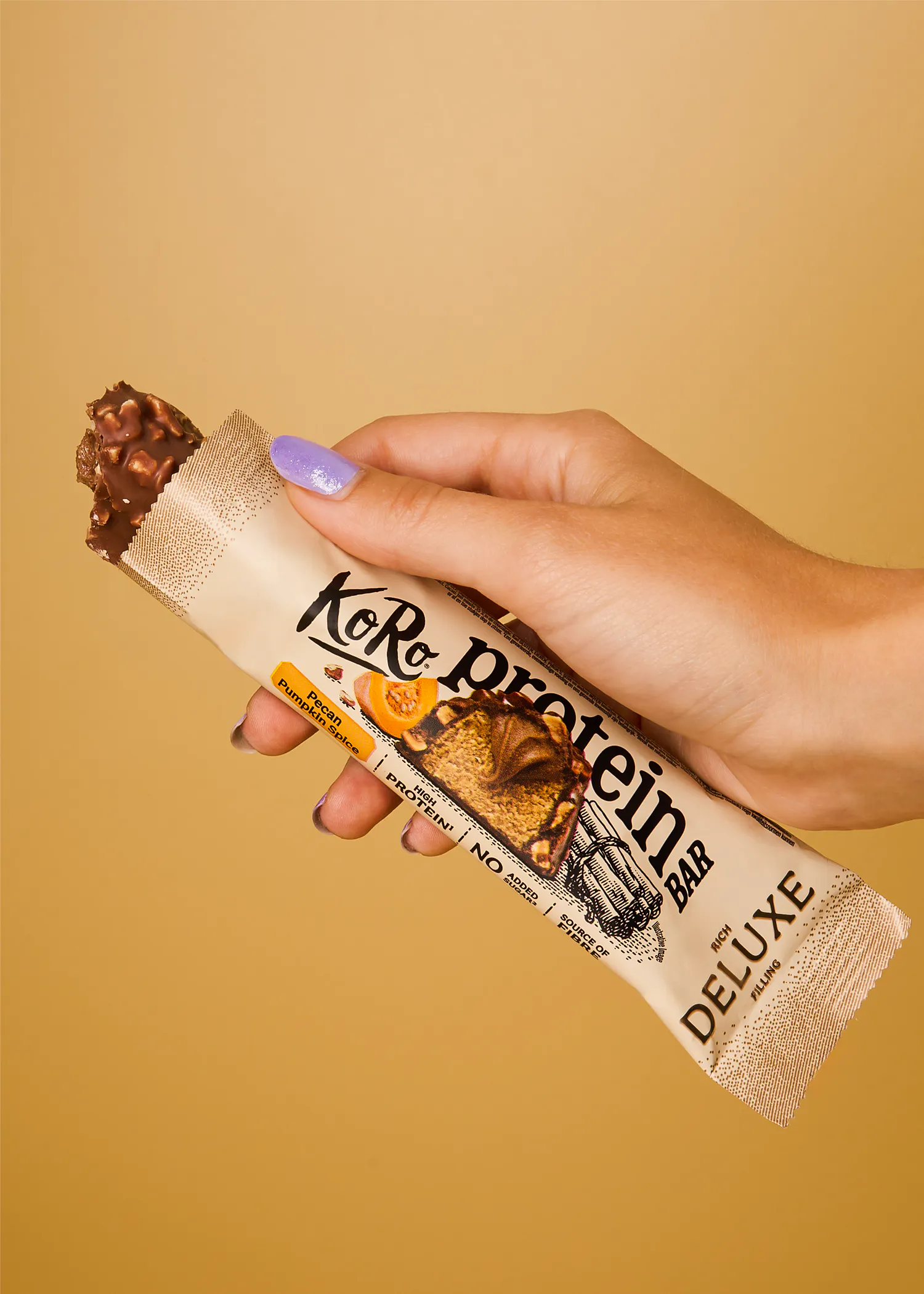 eine hand mit hellvioletten fingernägeln hält einen koro proteinriegel der sorte pecan pumpkin spice vor einem beigen hintergrund.