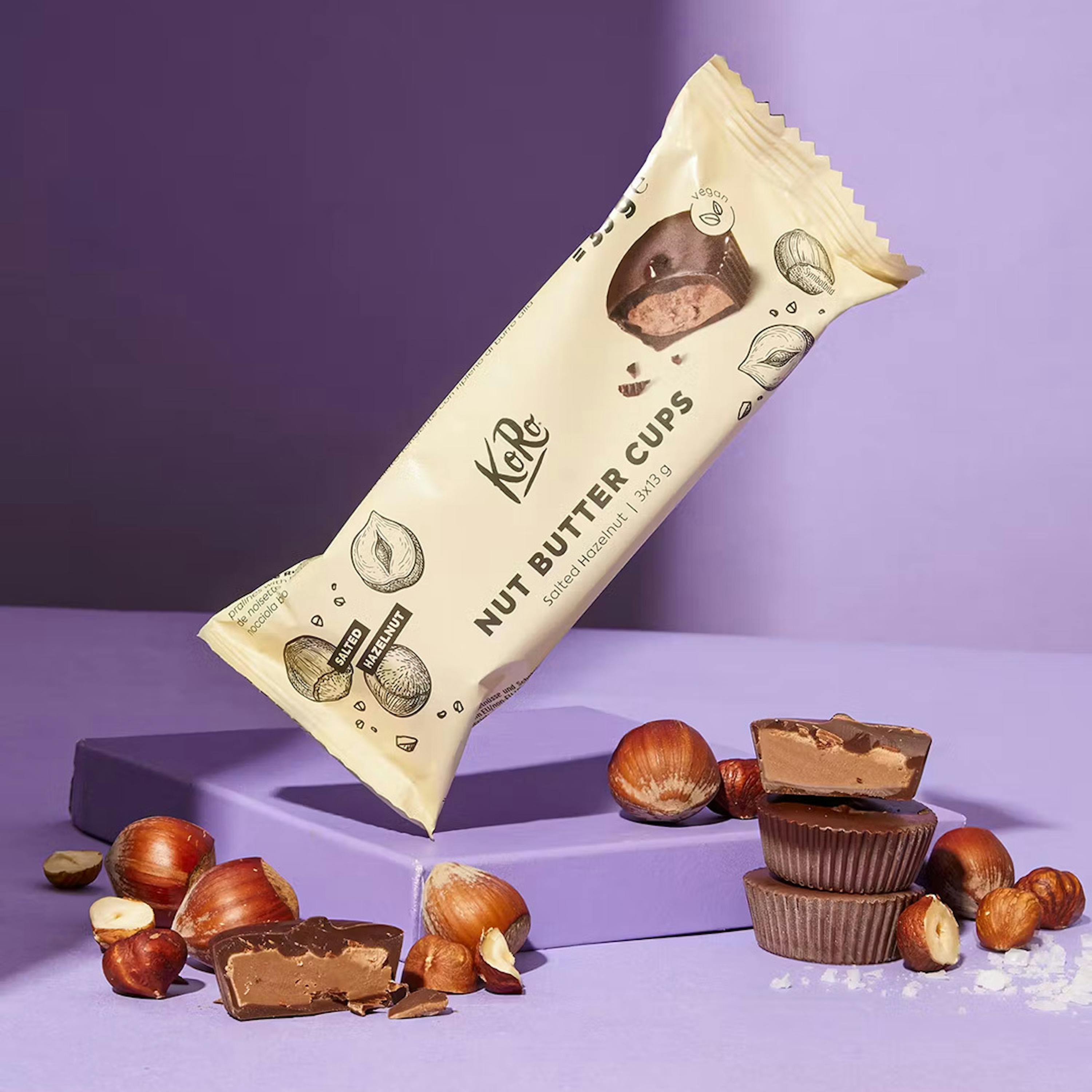 un emballage de ko ro nut butter cups à la noisette salée flottant au-dessus d'une plateforme violette, entouré de noisettes et de tasses de chocolat coupées en deux révélant leur garniture.