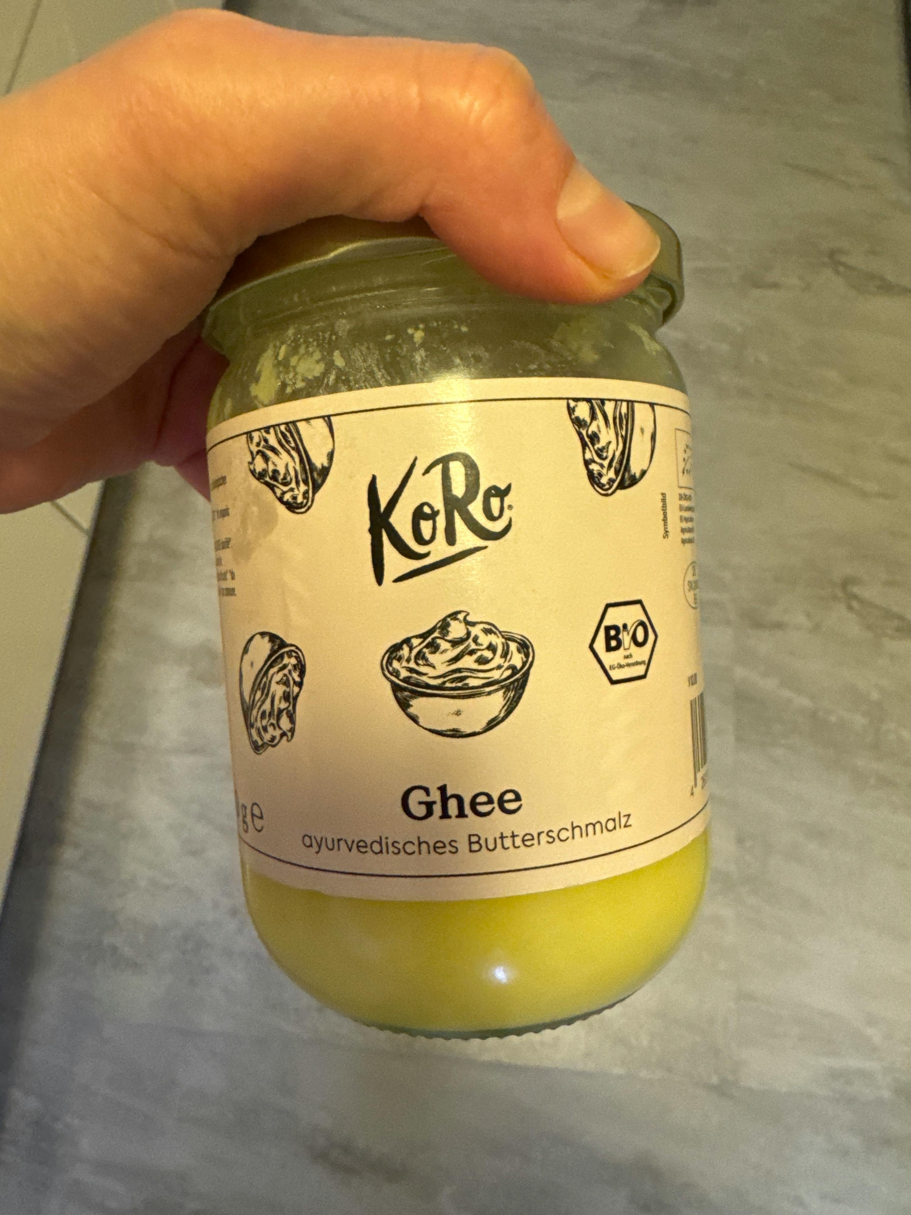 eine hand hält ein glasgefäß mit kero ghee, einem ayurvedischen butterschmalz, mit einem bio-etikett.