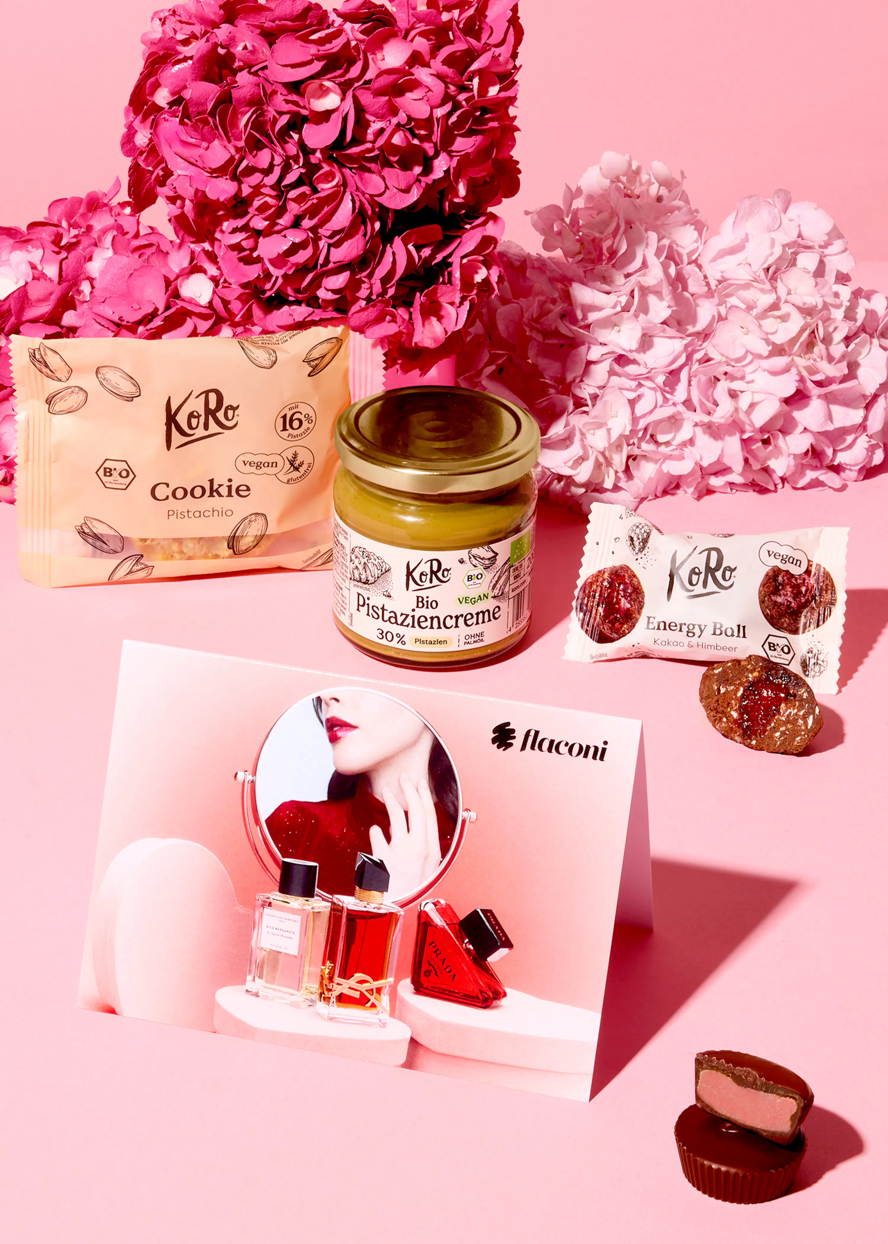 koro-produkte wie pistazienkekse, pistaziencreme und energiekugeln sind zusammen mit einem flaconi-display, das parfümflaschen und einen spiegel mit dem gesicht einer frau zeigt, auf einem rosa hintergrund mit hortensien arrangiert.
