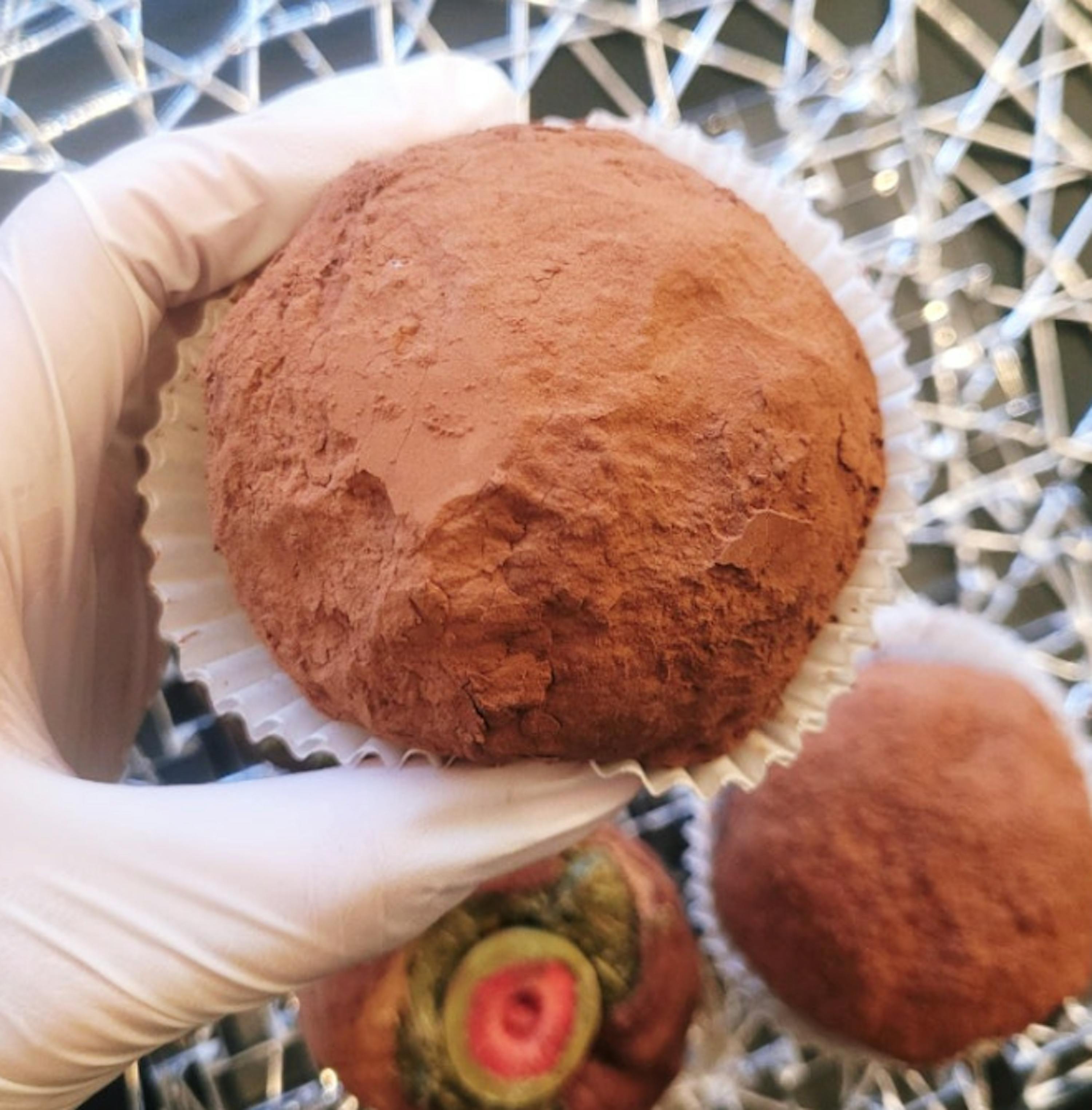 una mano guantata di bianco tiene un tartufo al cioccolato ricoperto di cacao in una coppetta di carta.