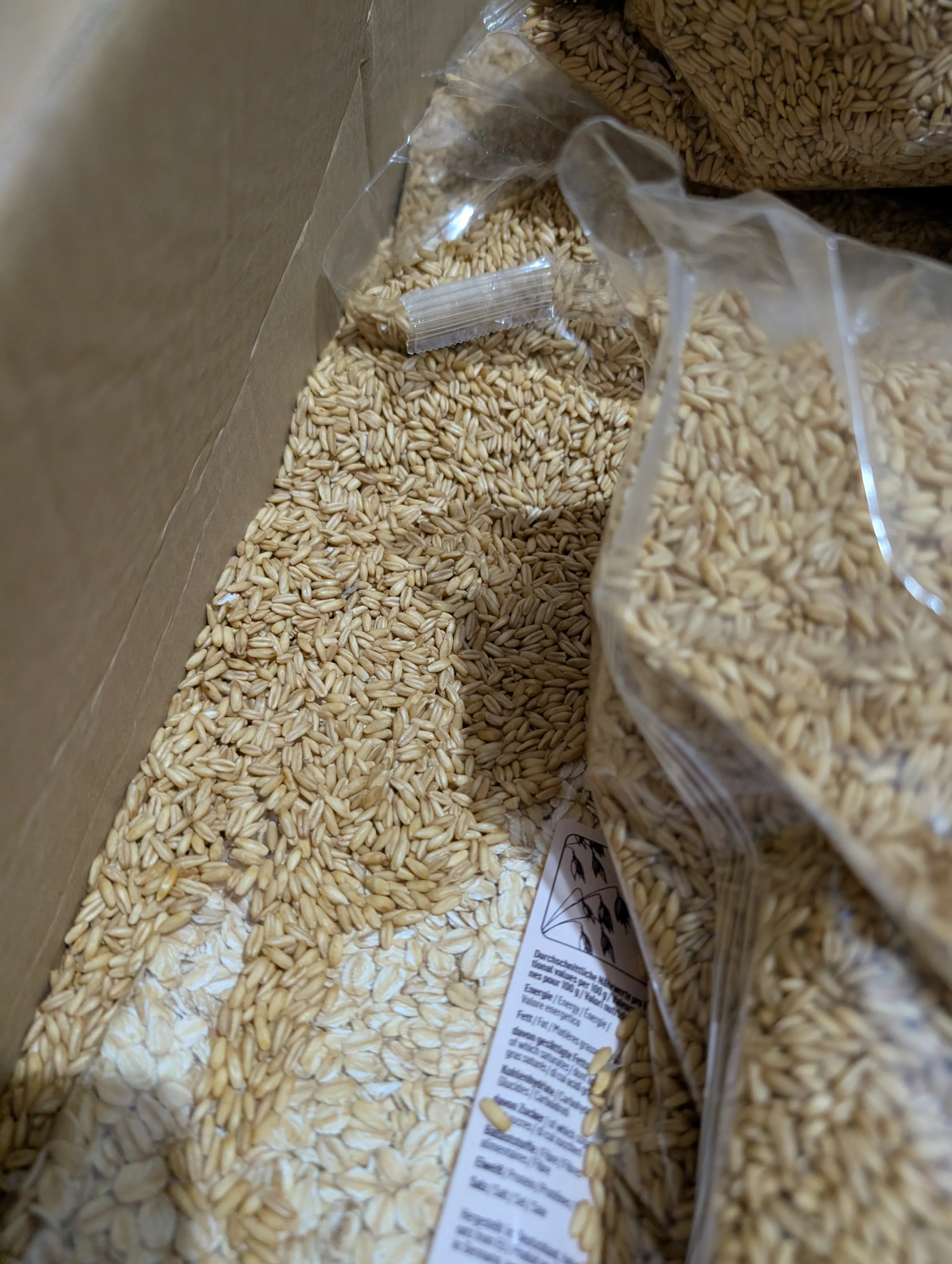una scatola di cartone piena di chicchi d'avena sfusi e in sacchetti di plastica, con un'etichetta nutrizionale parzialmente visibile.