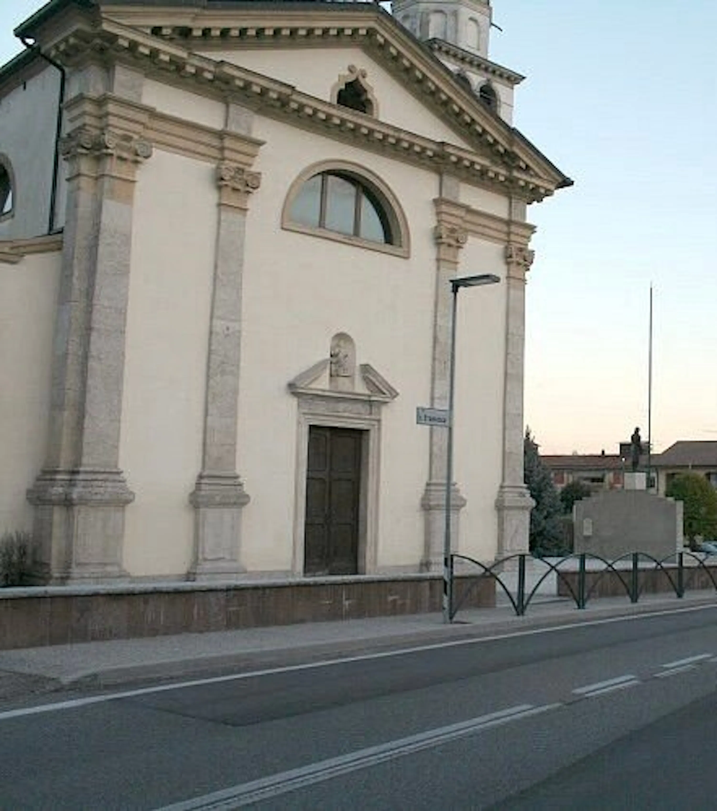 facciata di una chiesa color crema con colonne, un portale decorato e una finestra ad arco, affacciata su una strada con una recinzione metallica.