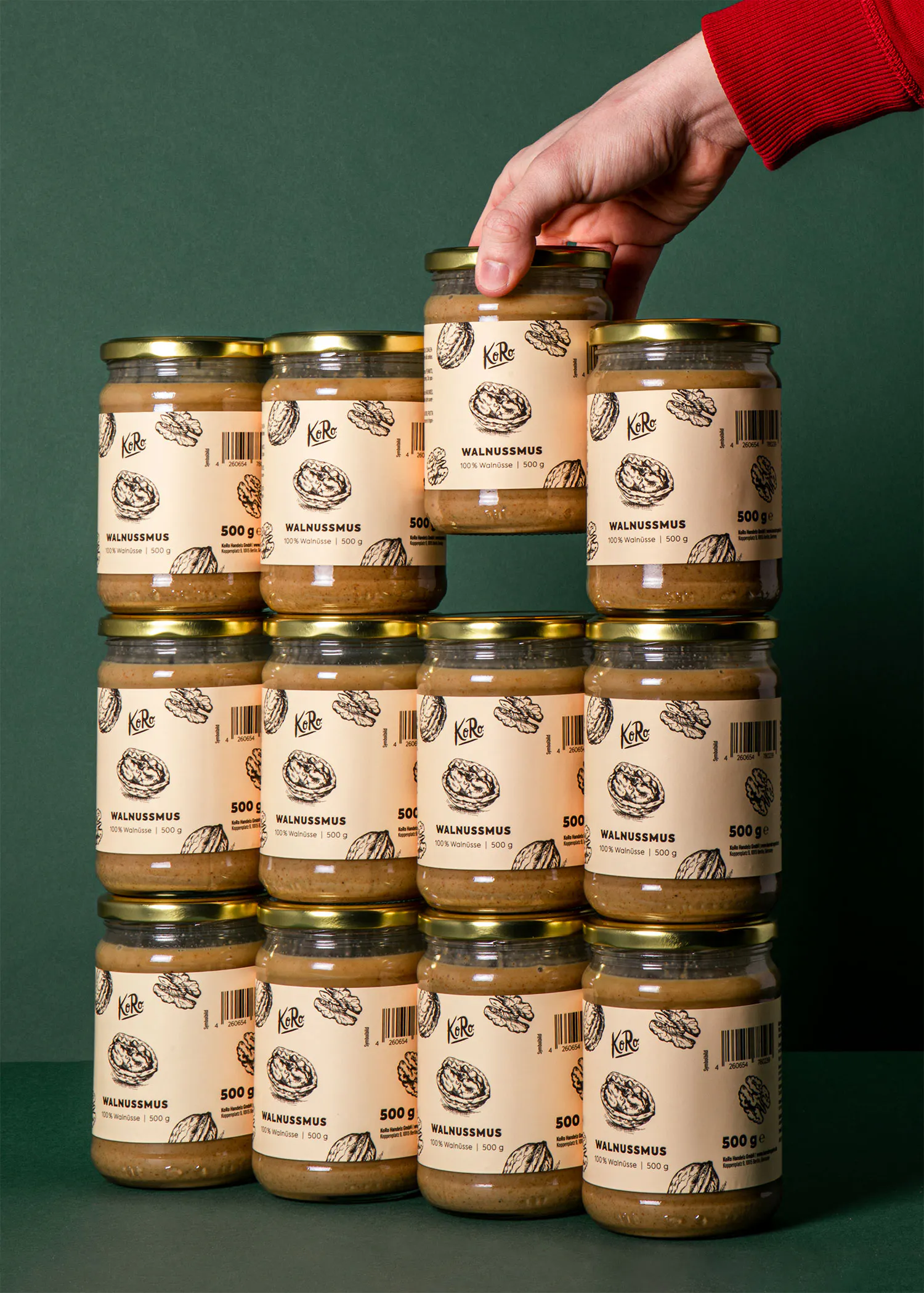 une main tient un pot de purée de noix koro au sommet d'une pile de douze pots identiques disposés sur un fond vert.
