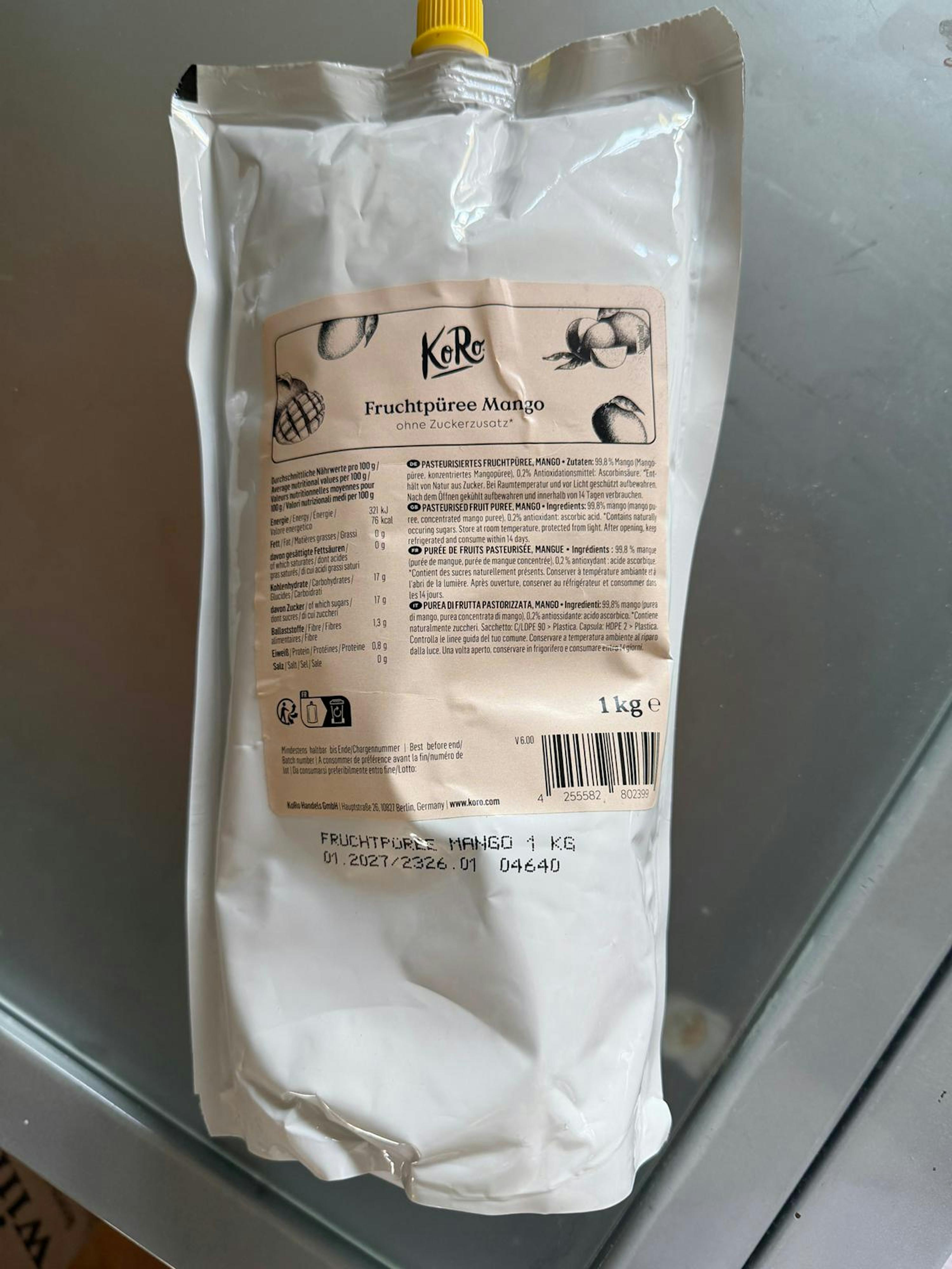 a white koro mango fruit puree pouch with a yellow cap, featuring a label with mango illustrations, nutritional information, and text for 1 kg and no added sugar.