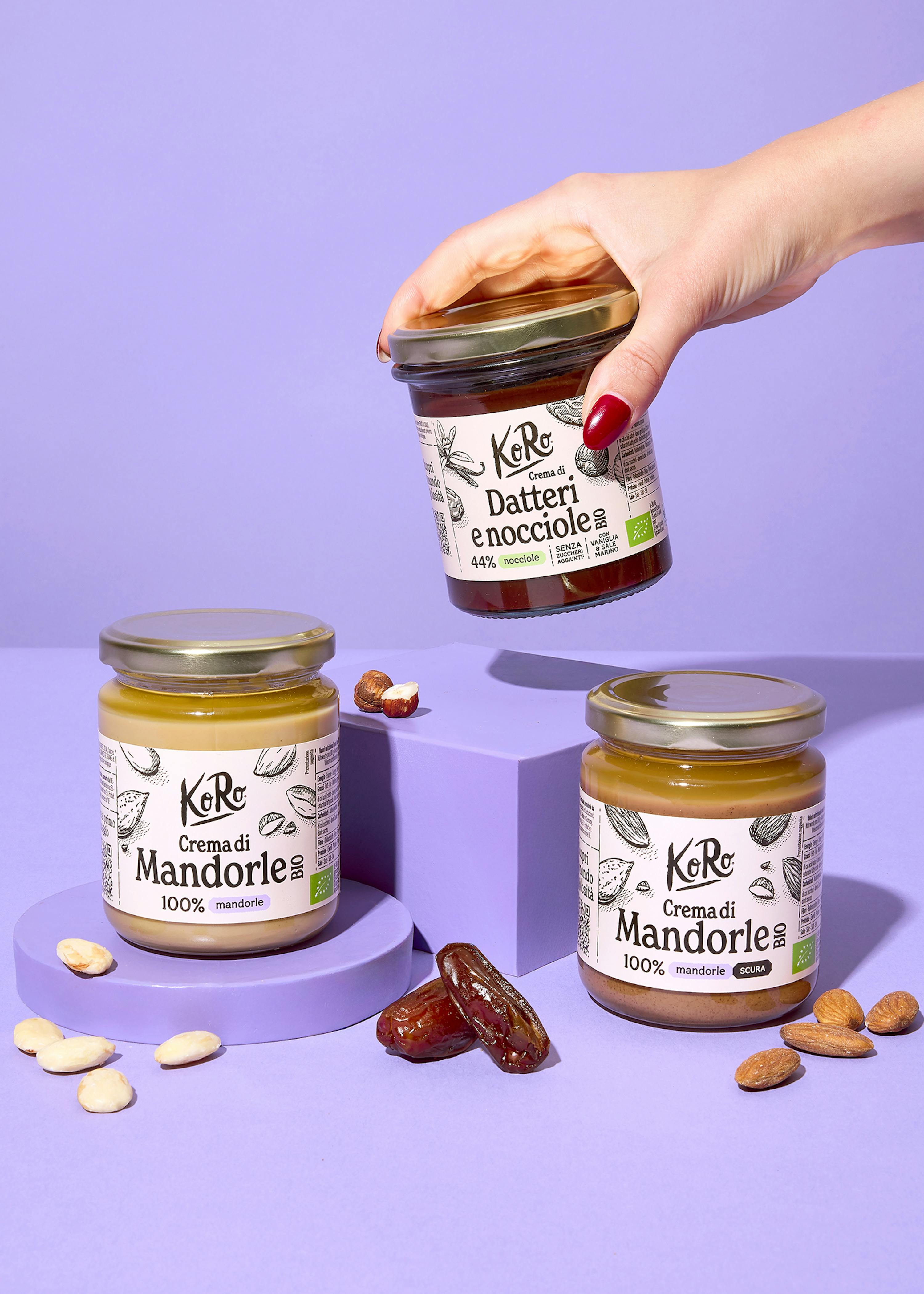una mano con smalto rosso tiene un vasetto di crema di datteri e nocciole koro, mentre altri due vasetti di crema di mandorle koro, uno chiaro e uno scuro, sono appoggiati su blocchi viola, circondati da datteri, nocciole e mandorle sparse.