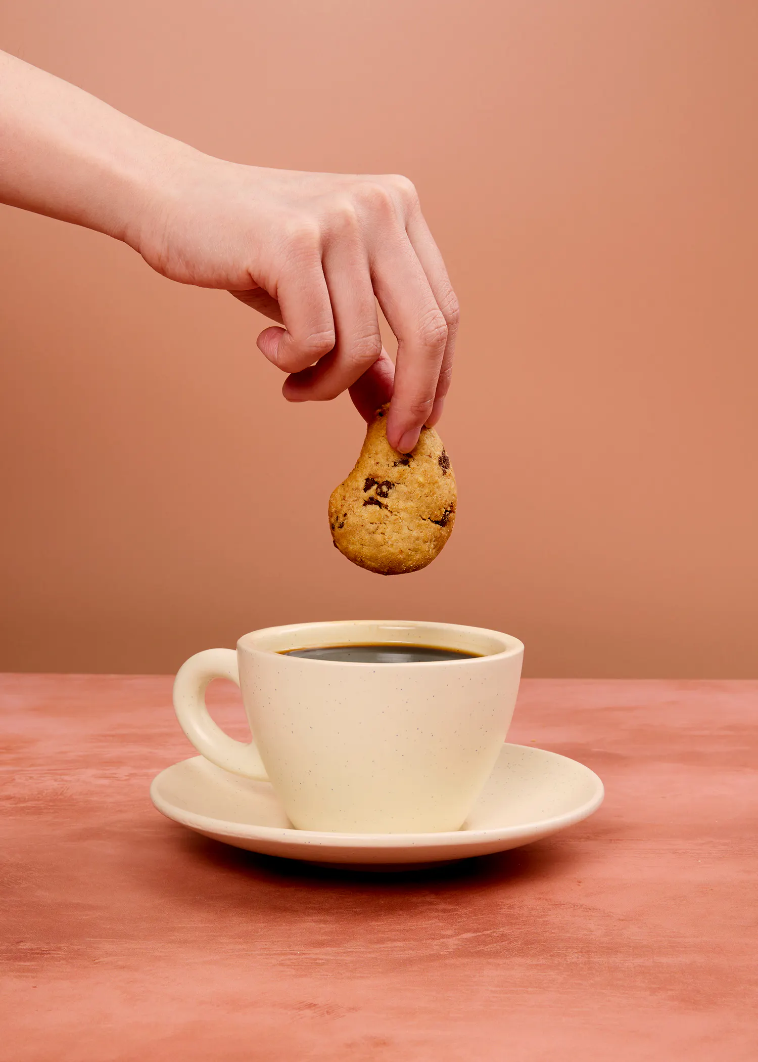 una mano sostiene una galleta con chispas de chocolate sobre una taza de café.