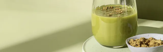 ein glas mit grünem smoothie steht neben einer kleinen schale mit grünem pulver auf einem hellgrünen untergrund.