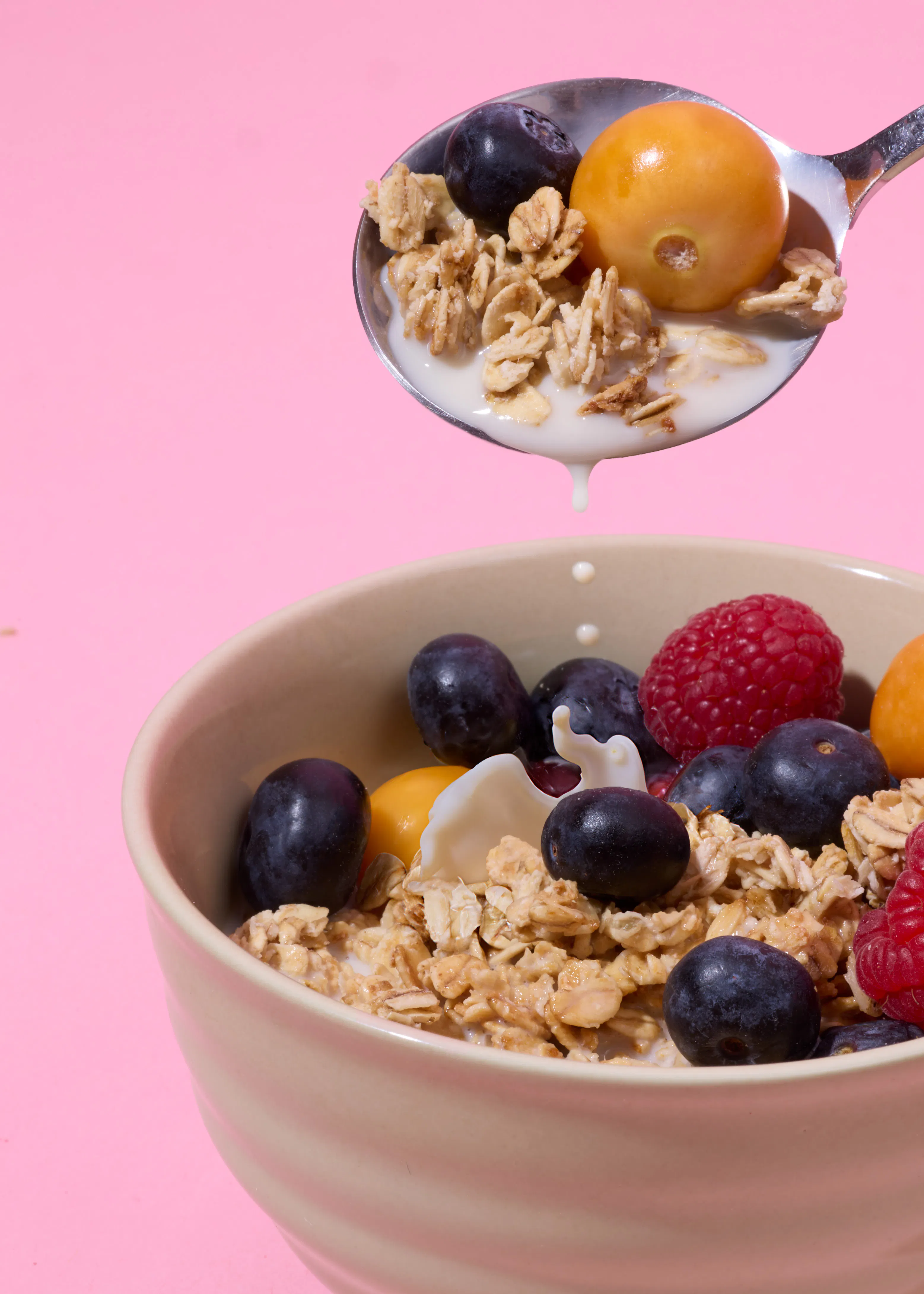 a spoon filled with granola, a blueberry, and a goldenberry drips milk into a bowl of granola, blueberries, raspberries, and goldenberries, all against a pink background.