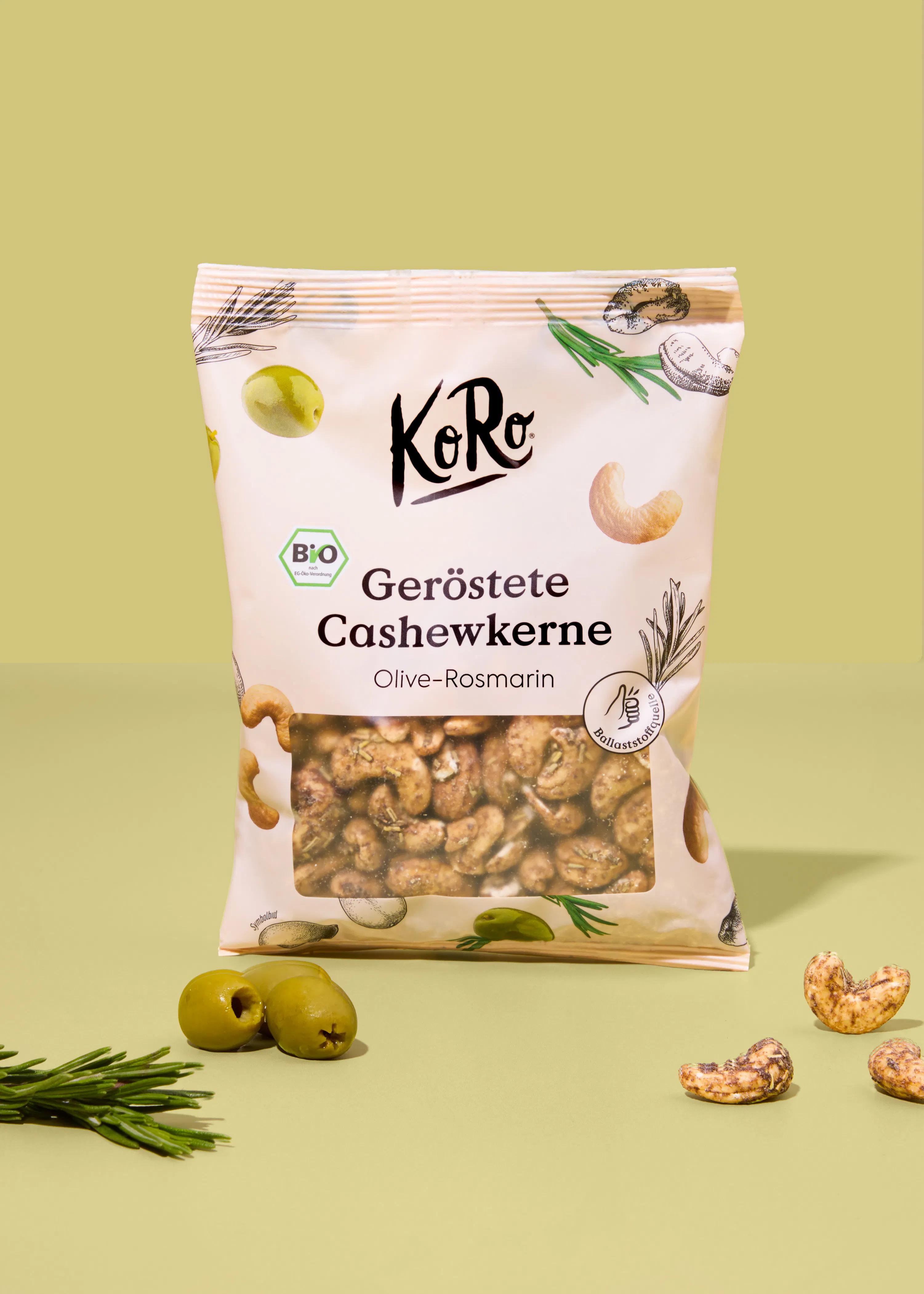 confezione di anacardi tostati al gusto di oliva e rosmarino del marchio koro su sfondo verde chiaro con olive e rametti di rosmarino freschi.