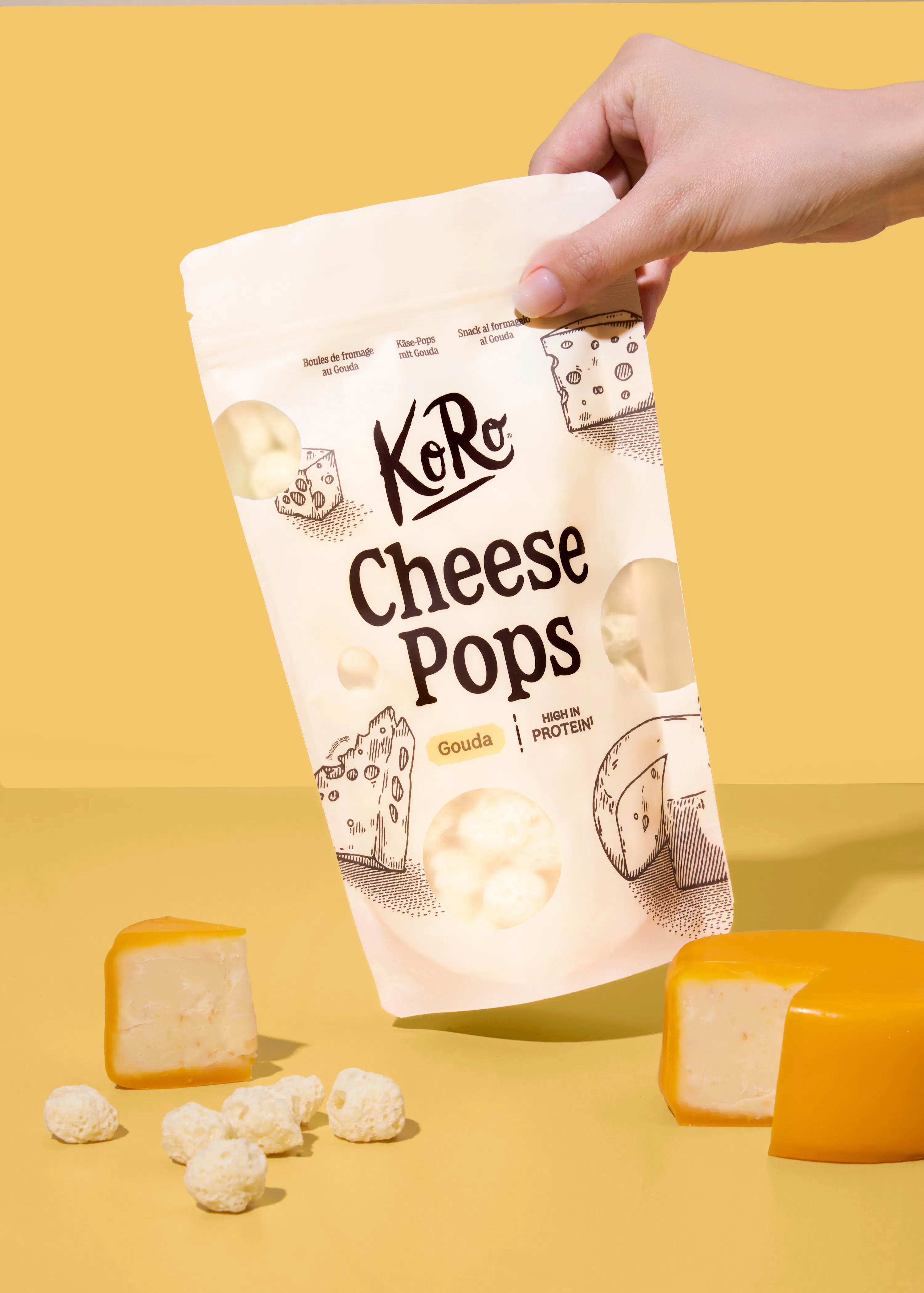 una mano tiene una confezione di koro cheese pops al gouda su uno sfondo giallo, con pezzi di formaggio gouda e snack croccanti sparsi sul piano.