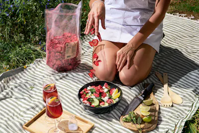 una mujer se sienta en una manta junto a un tazón de ensalada de fresas y pepinos