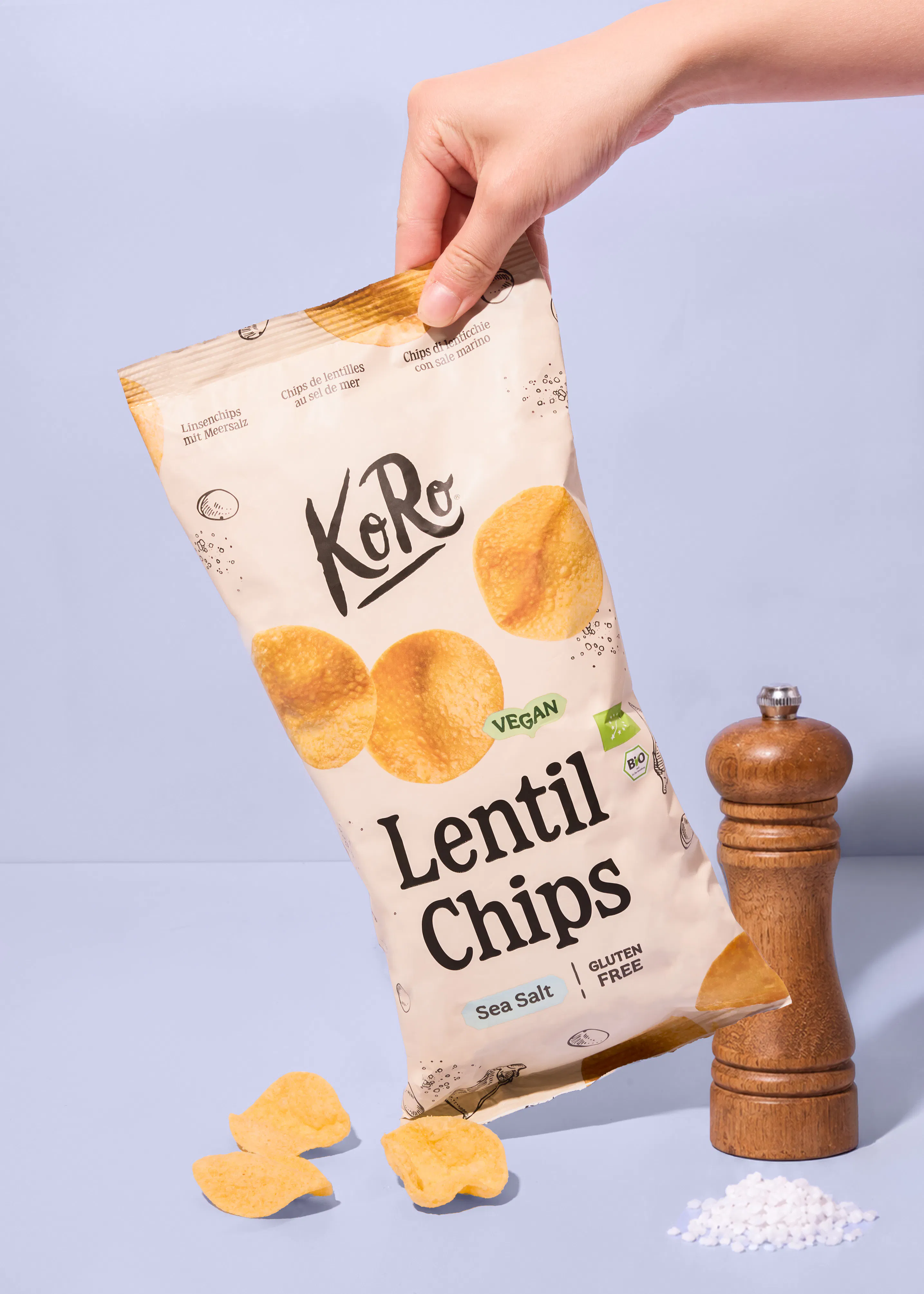 une main tient un paquet de chips de lentilles koro au sel de mer au-dessus d'un moulin à poivre et de quelques chips éparpillées sur une surface bleue.