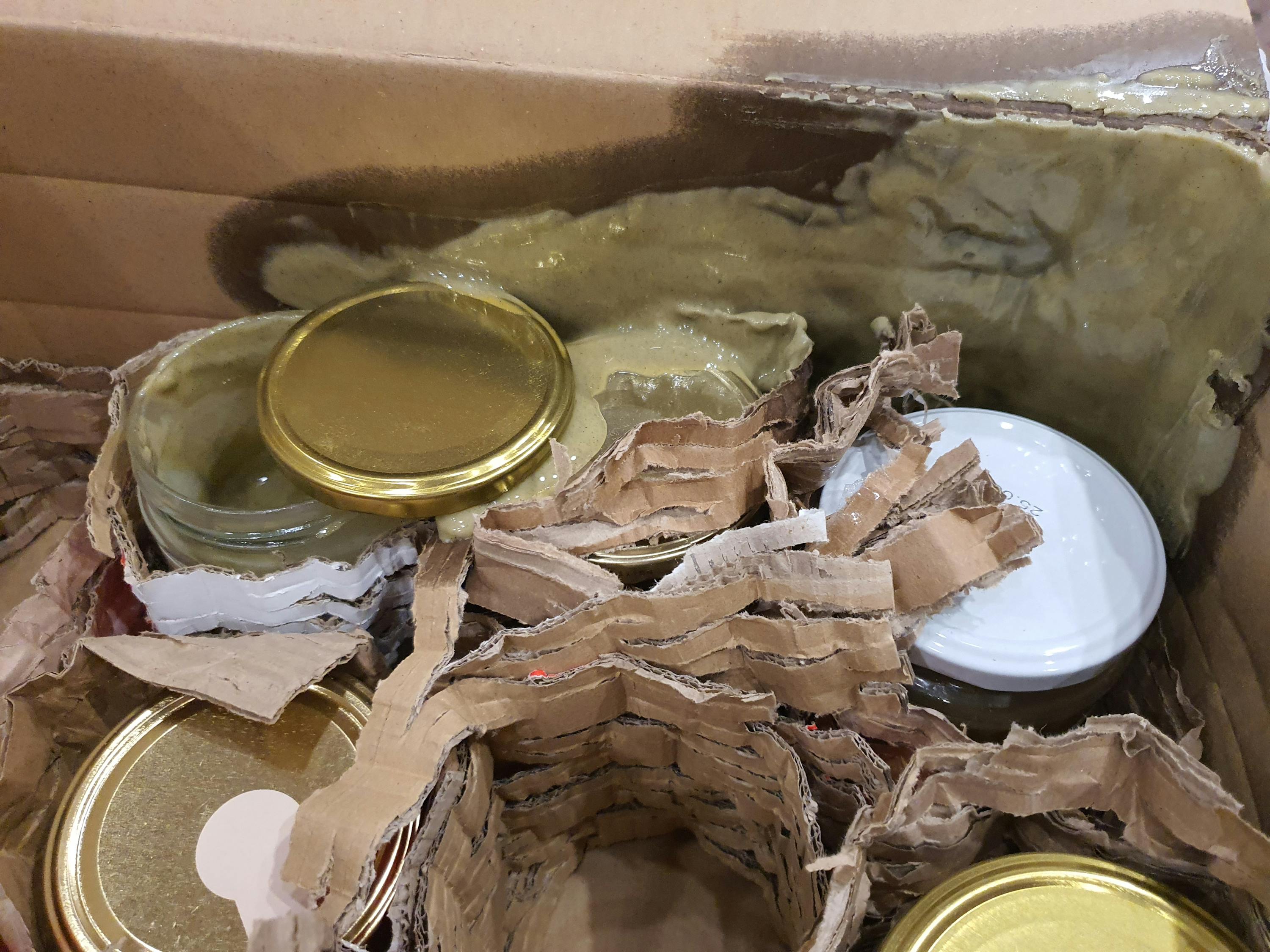 eine braune versandbox mit mehreren gläsern, die mit wellpappe getrennt sind, wobei eine grüngraue substanz aus einem der gläser ausgelaufen ist und die verpackung verschmiert.