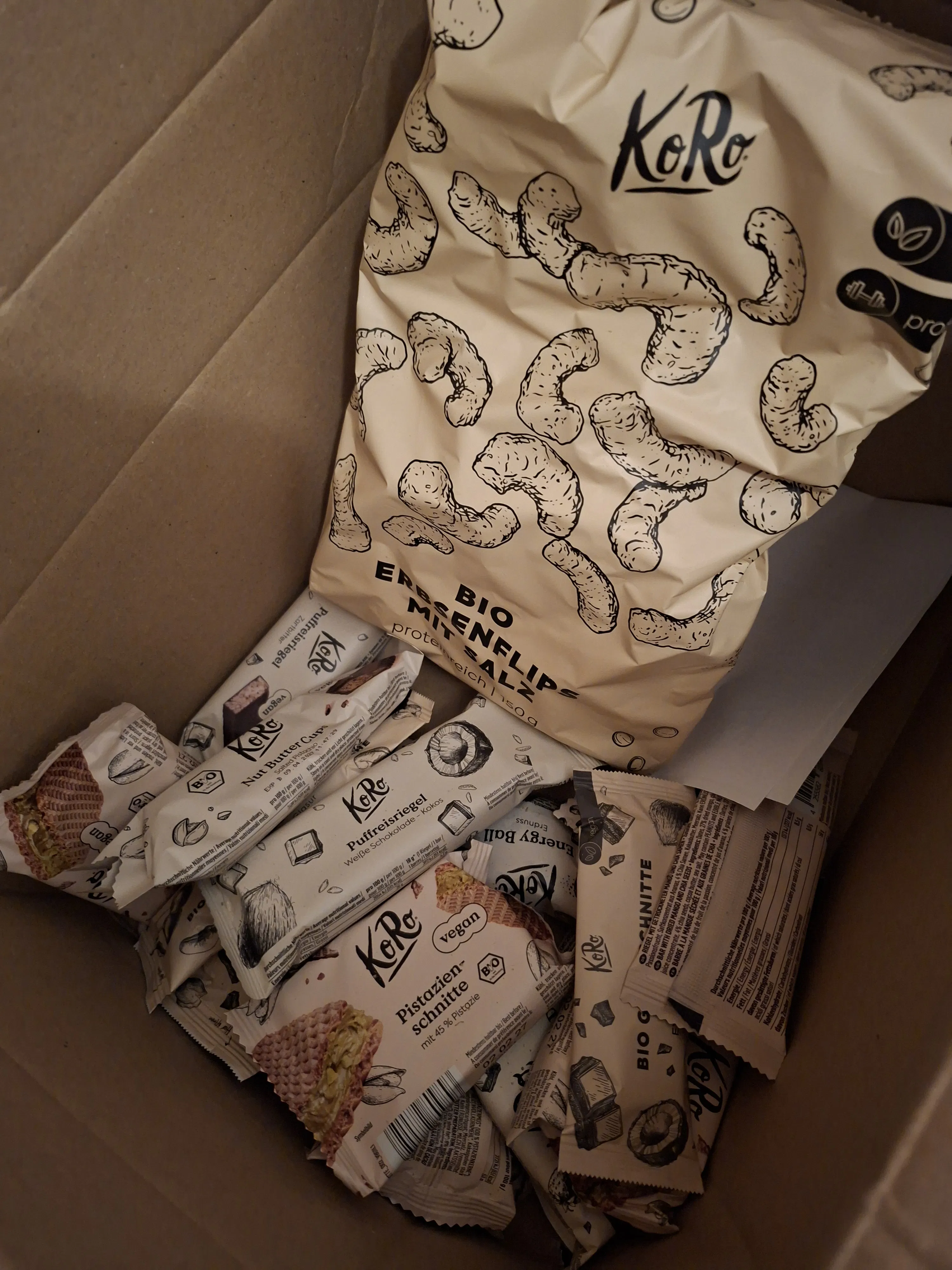 eine offene pappschachtel gefüllt mit verschiedenen koro-snacks, darunter eine tüte bio-erbsenflips mit salz und mehrere einzeln verpackte riegel wie pistazien-schnitten und puffreisriegel.