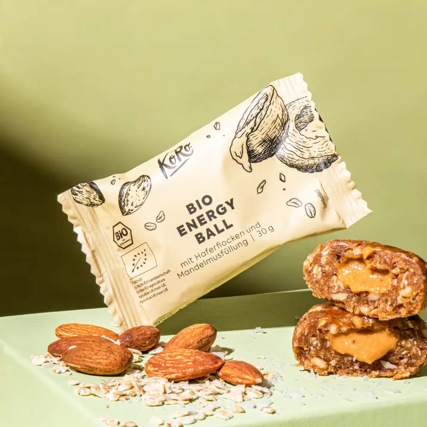 un sachet de bio energy ball koro à l'avoine et au beurre d'amande posé à côté d'amandes entières et d'une boule énergétique coupée en deux révélant son cœur fondant.