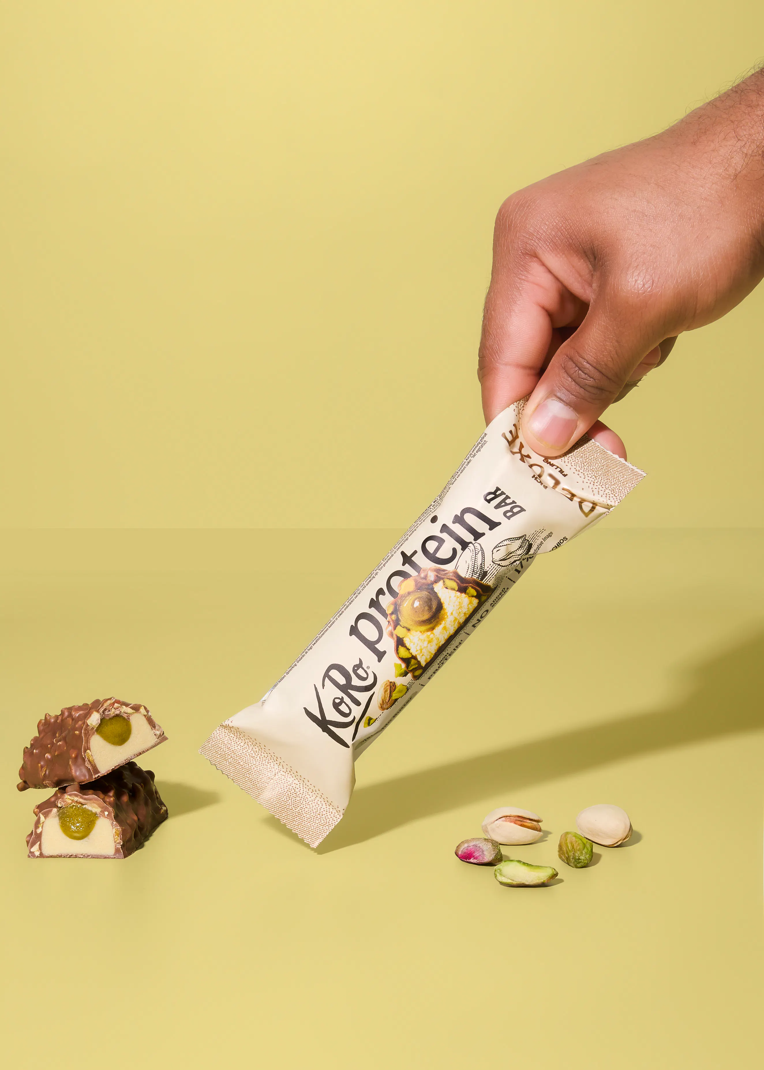 eine hand hält einen koro proteinriegel mit pistazienfüllung vor einem hellgelben hintergrund, wobei daneben zwei aufgeschnittene riegelstücke und einige pistazien liegen.