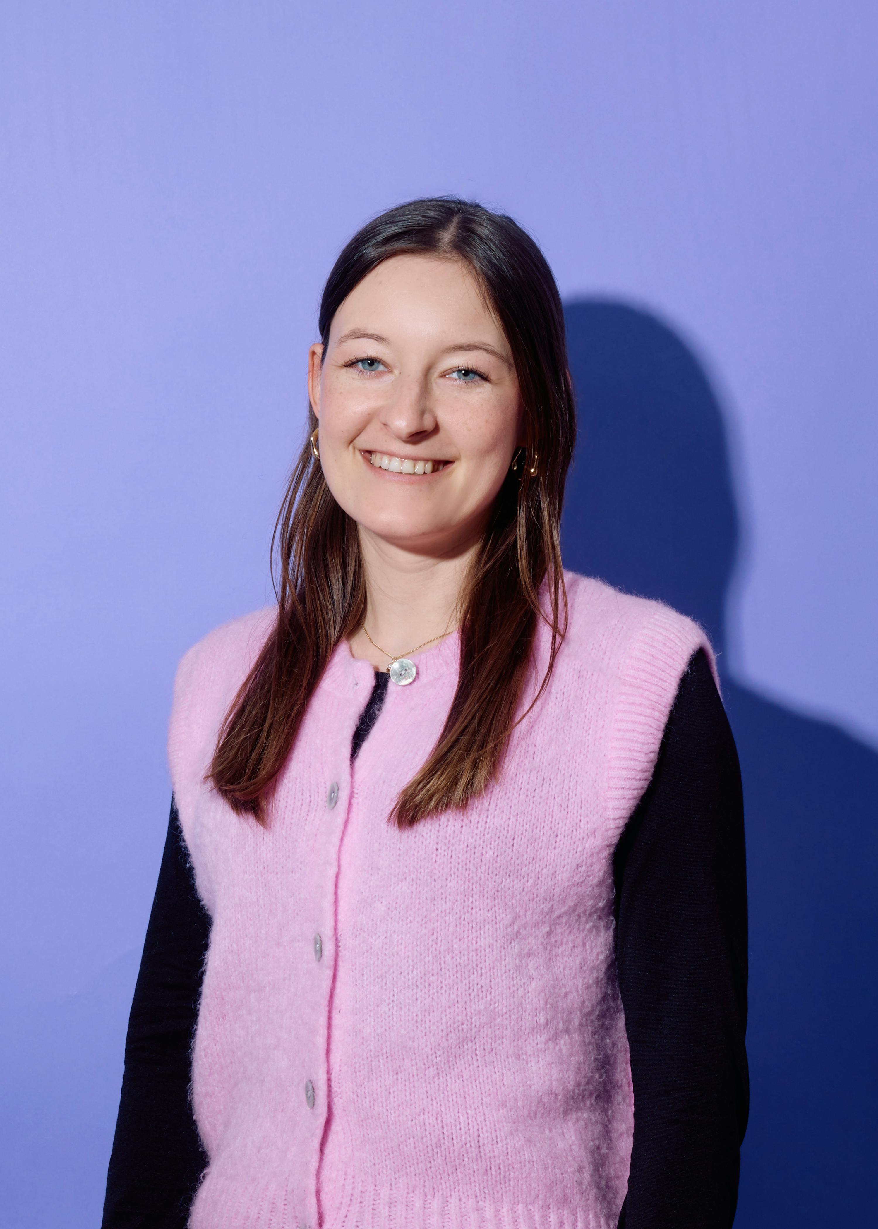 eine lächelnde junge frau mit langen braunen haaren, blauen augen und sommersprossen trägt eine rosa strickweste über einem schwarzen hemd vor einem lila hintergrund.