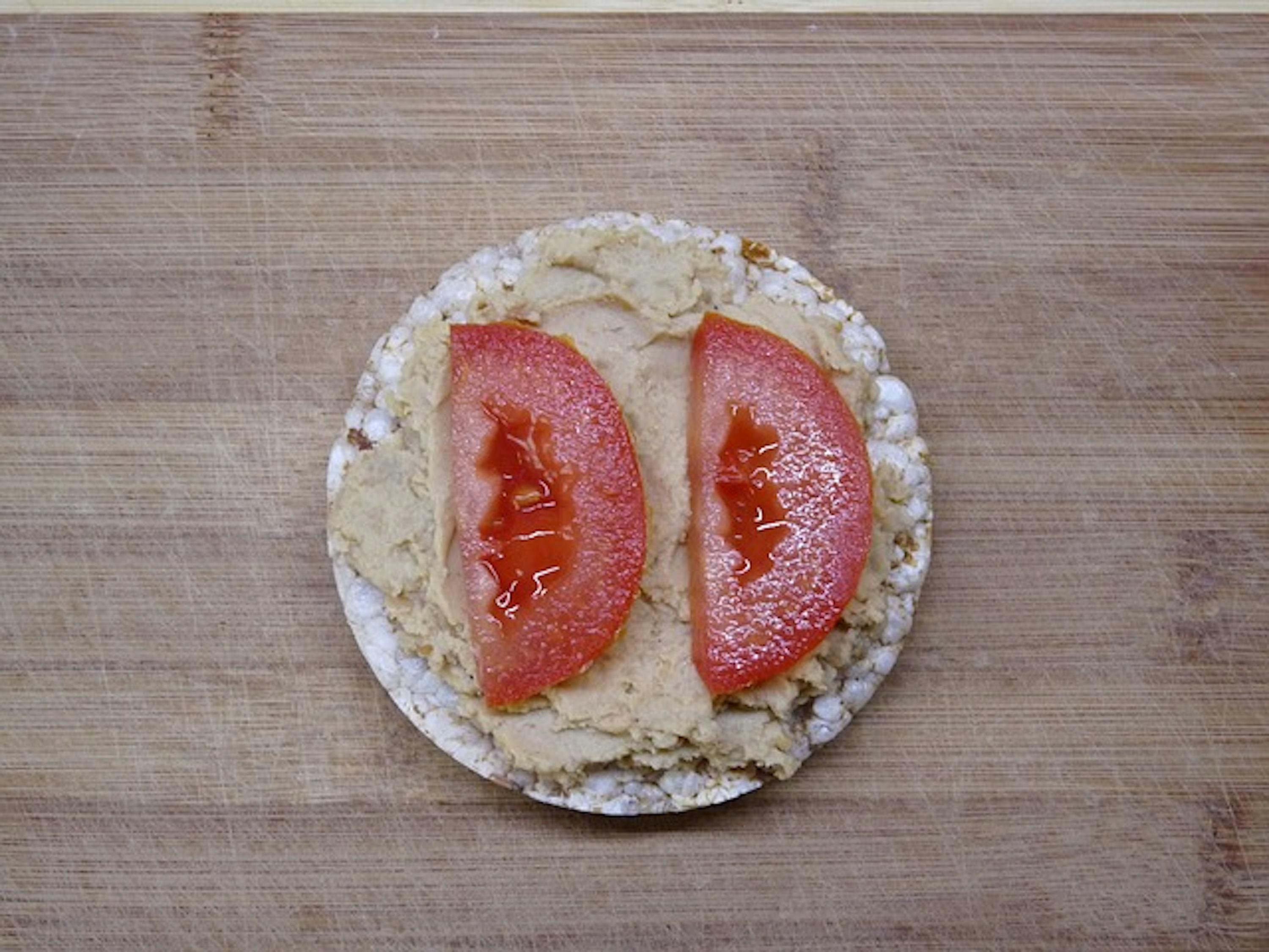 a rice cake with hummus and tomato slices on a wooden cutting board .
