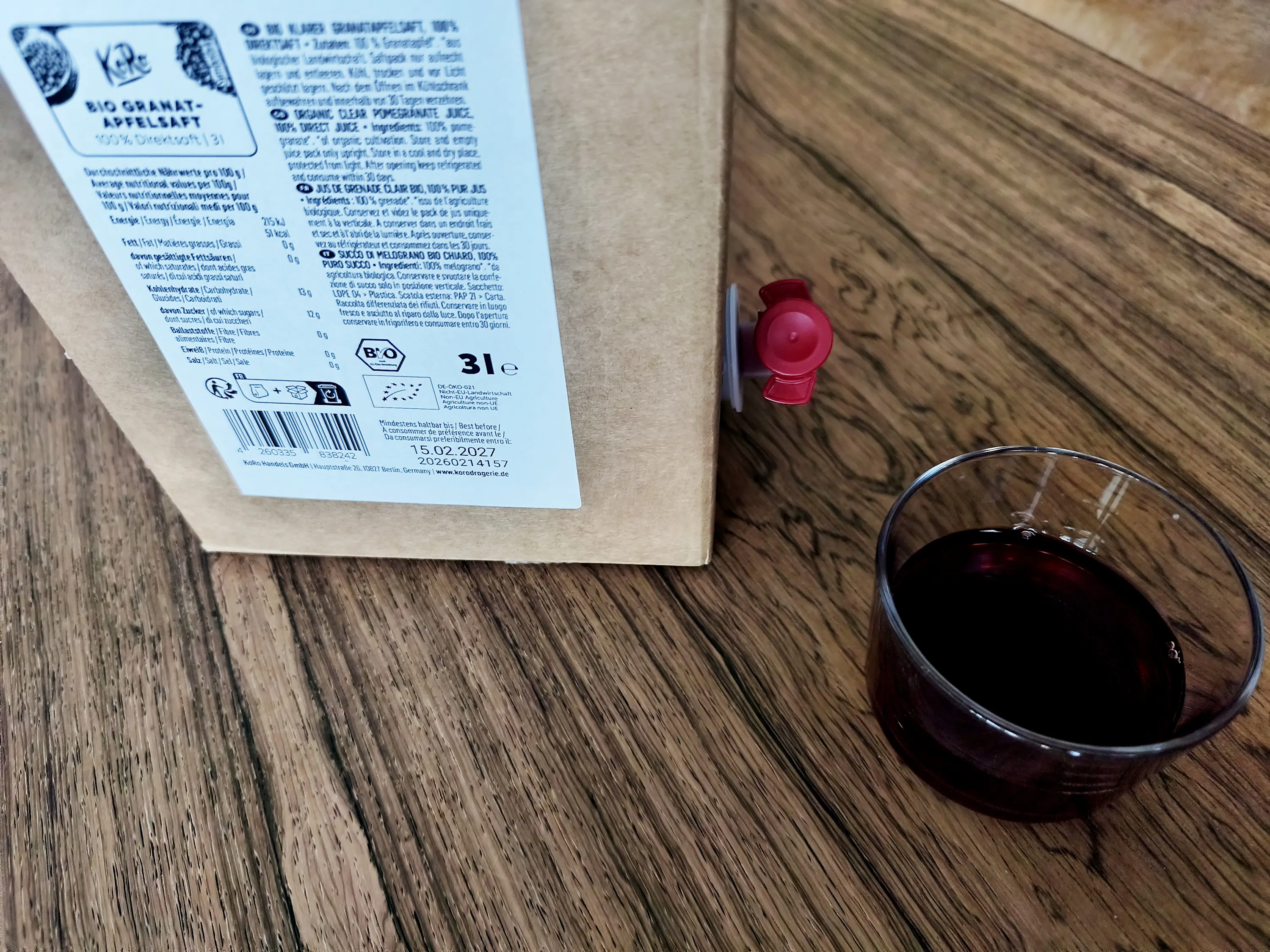 eine braune granatapfelsaftbox mit rotem zapfhahn steht auf einem holztisch neben einem glas mit dunklem saft.