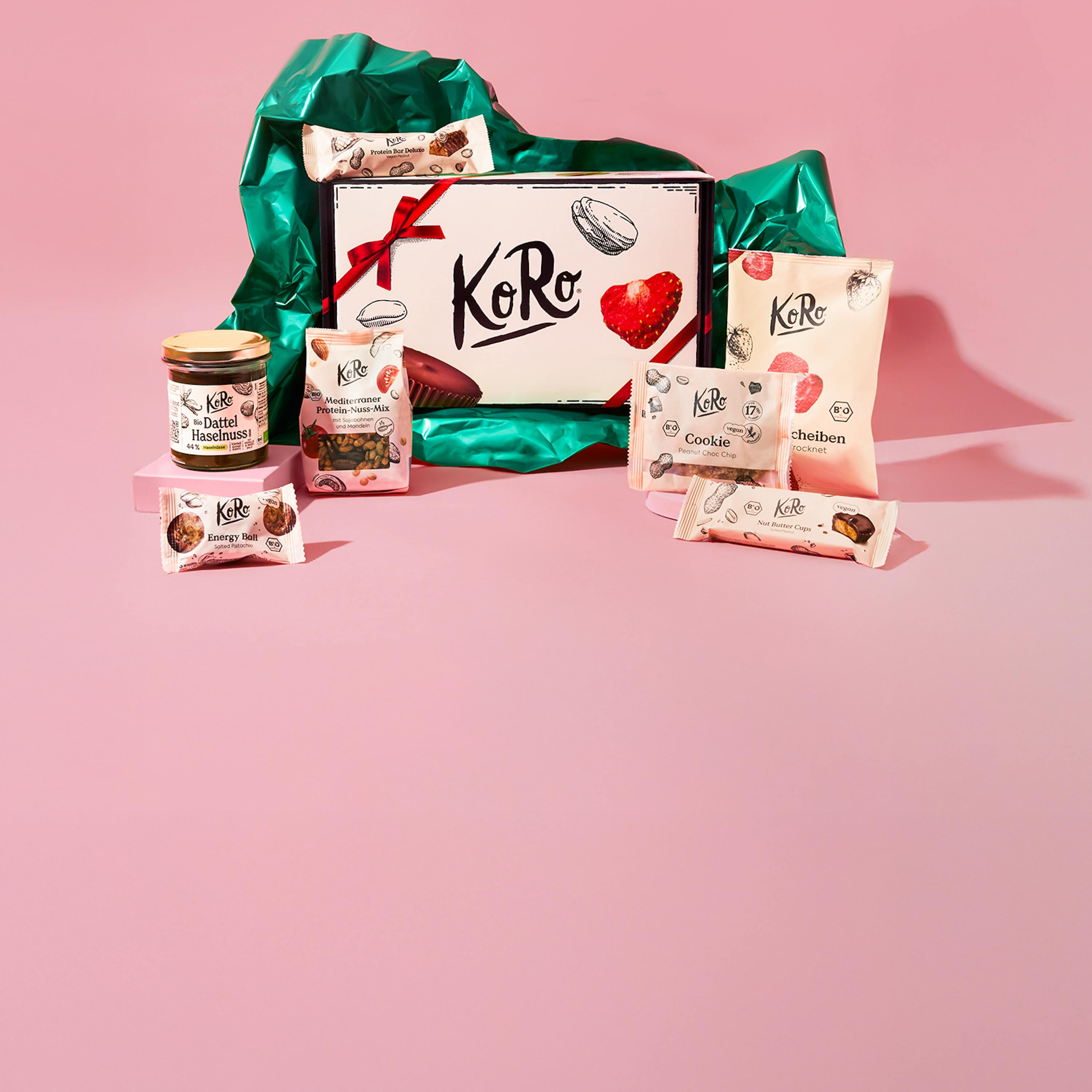 eine geschenkbox von koro mit roter schleife, umgeben von grünem seidenpapier, und verschiedenen koro-snacks wie dattel-haselnuss-creme, nussmischungen, proteinriegeln, cookies und getrockneten früchten auf einem pinken hintergrund.