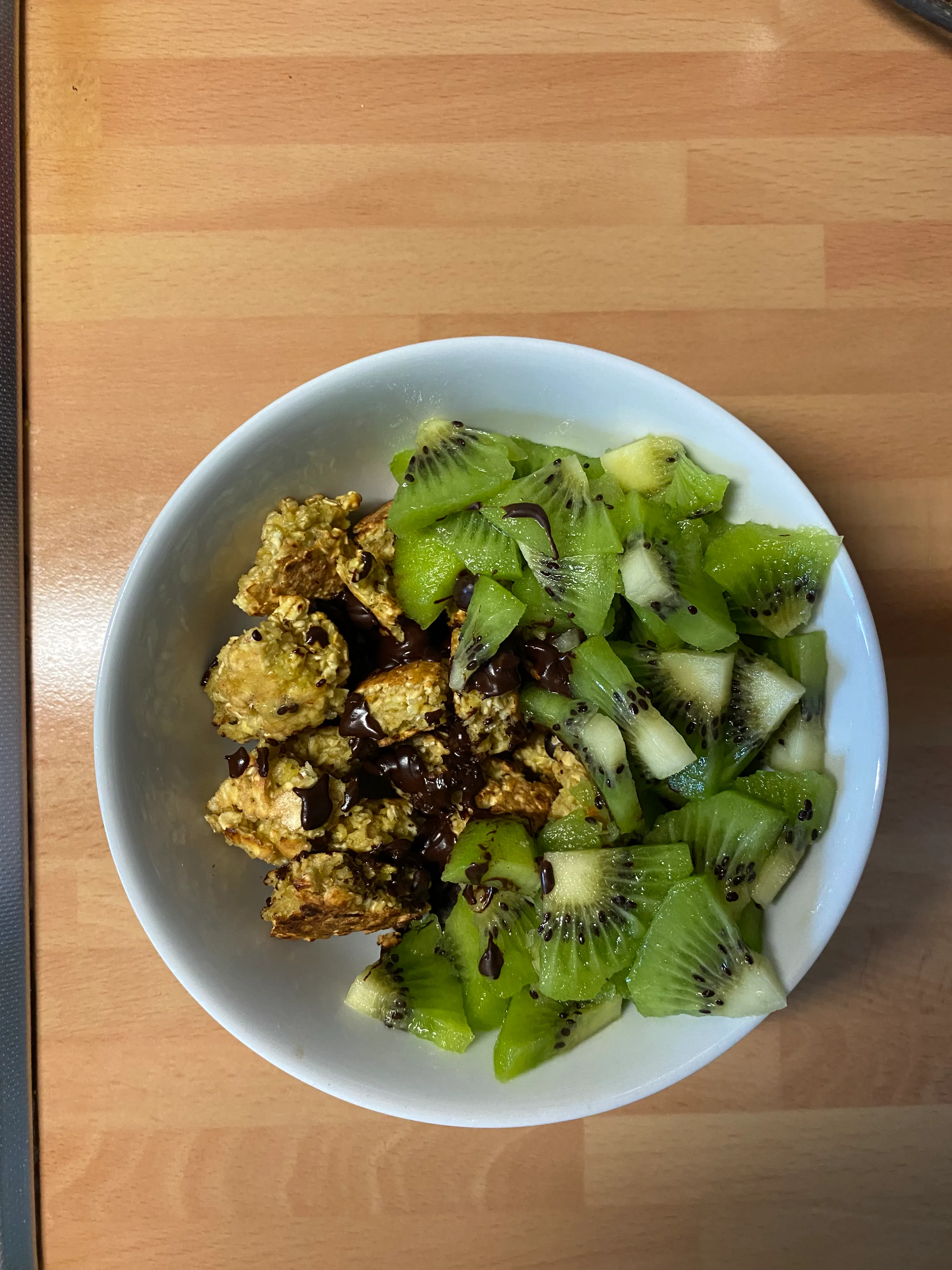 eine weiße schale gefüllt mit gewürfelten kiwis und gebackenen haferflocken, beträufelt mit schokolade, auf einem holztisch.