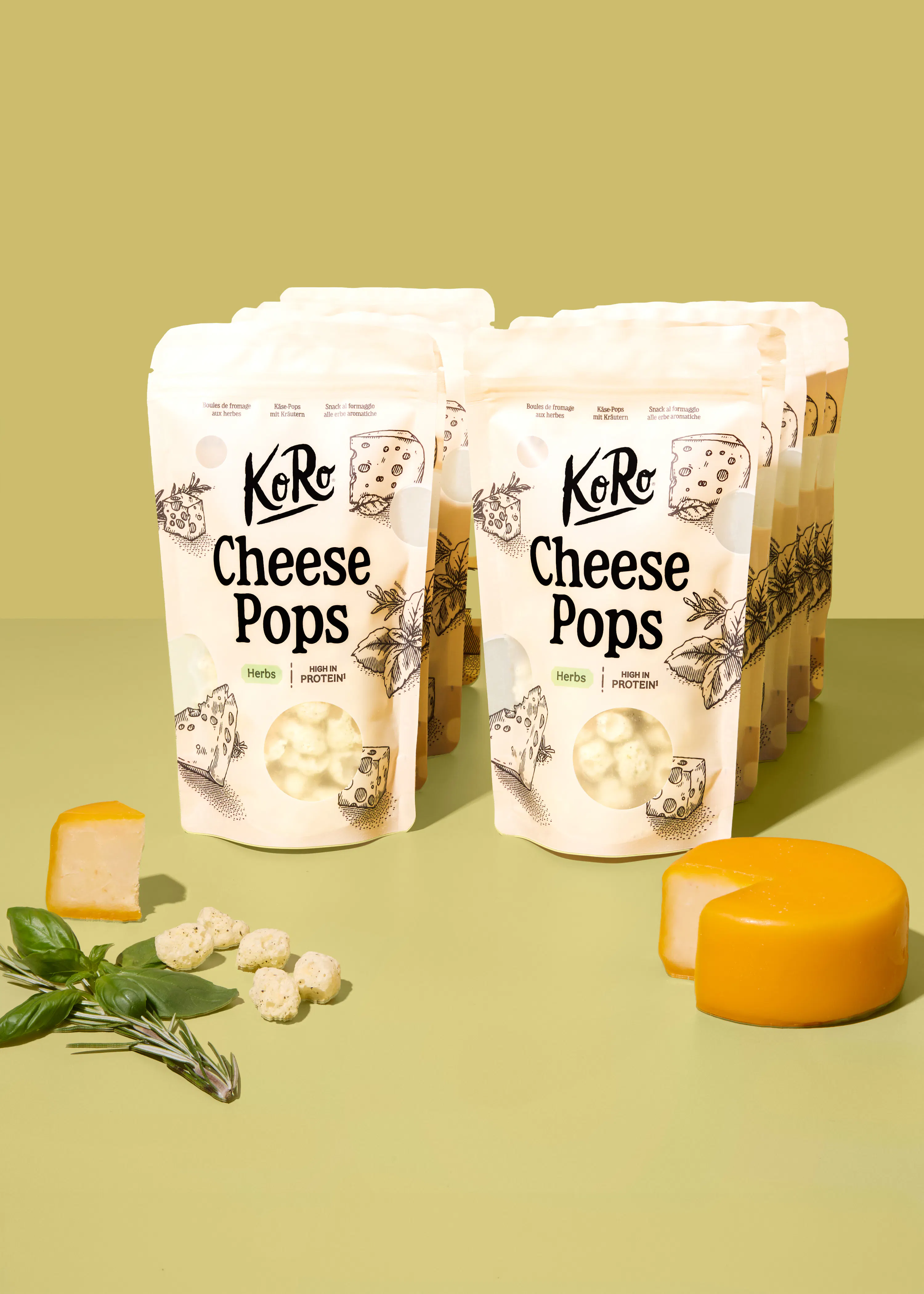 diverse confezioni di koro cheese pops alle erbe disposte su uno sfondo verde chiaro con accanto pezzi di formaggio e rametti di erbe aromatiche.