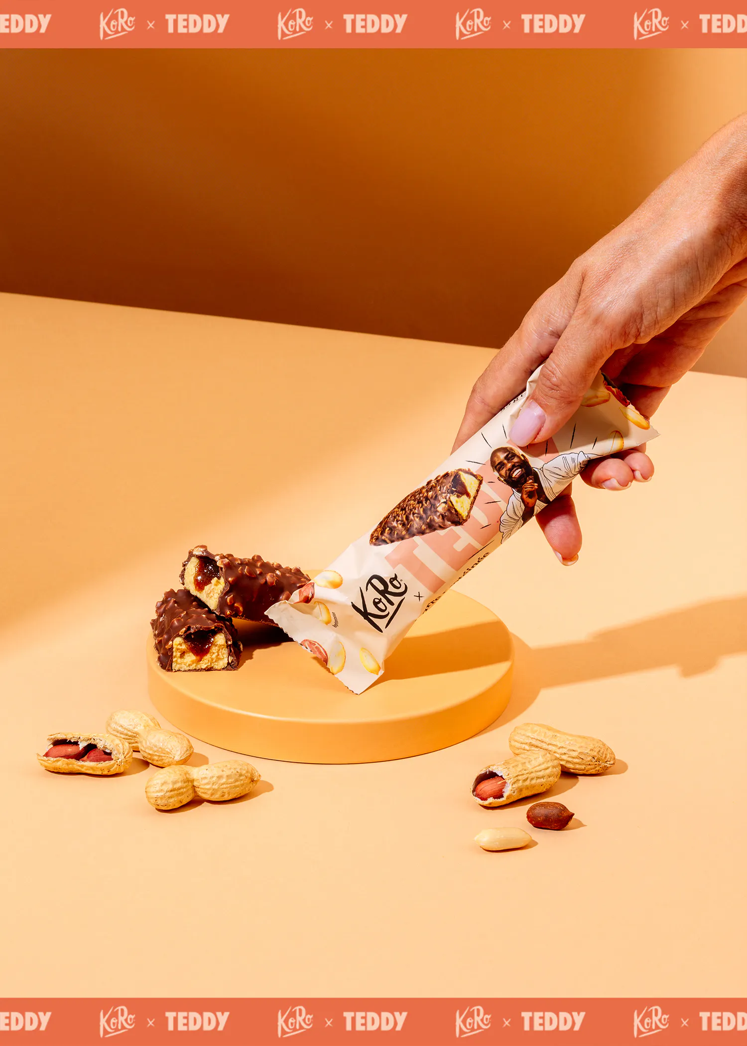 une main tient une barre chocolatée koro x teddy, avec des barres coupées et des cacahuètes éparpillées sur un fond orange.