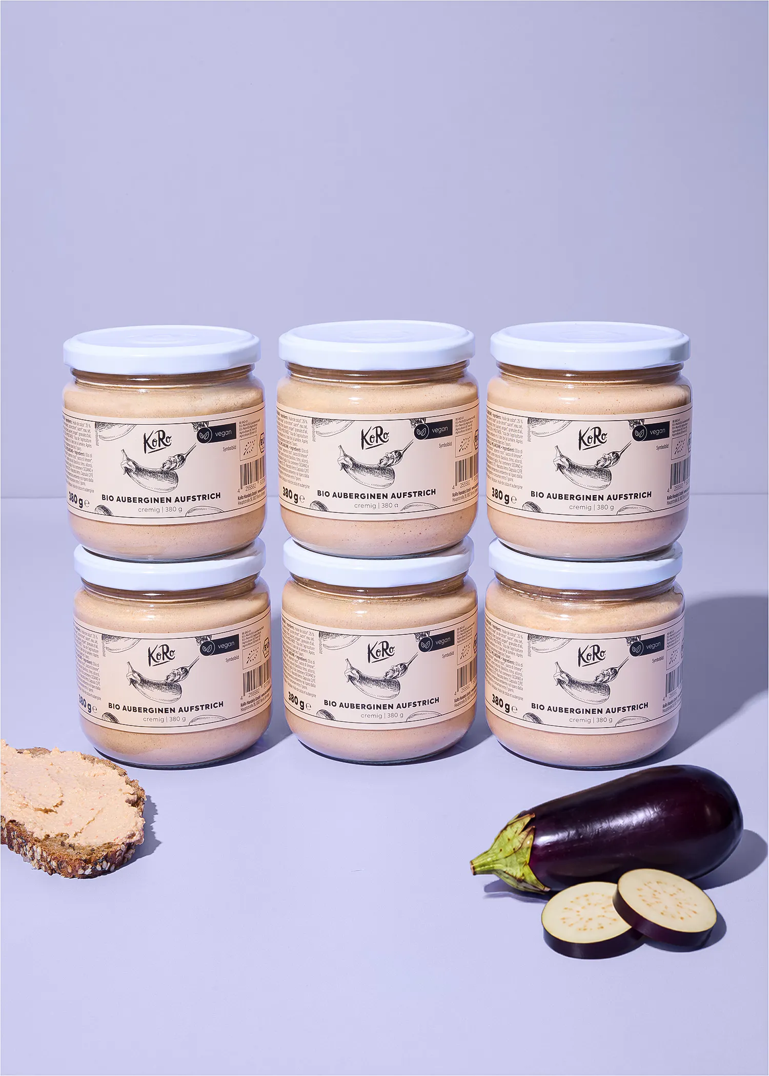 sechs gläser koro bio-auberginen-aufstrich sind in zwei reihen angeordnet, begleitet von einer scheibe brot mit aufstrich und einer frischen aubergine auf einem hellvioletten hintergrund.