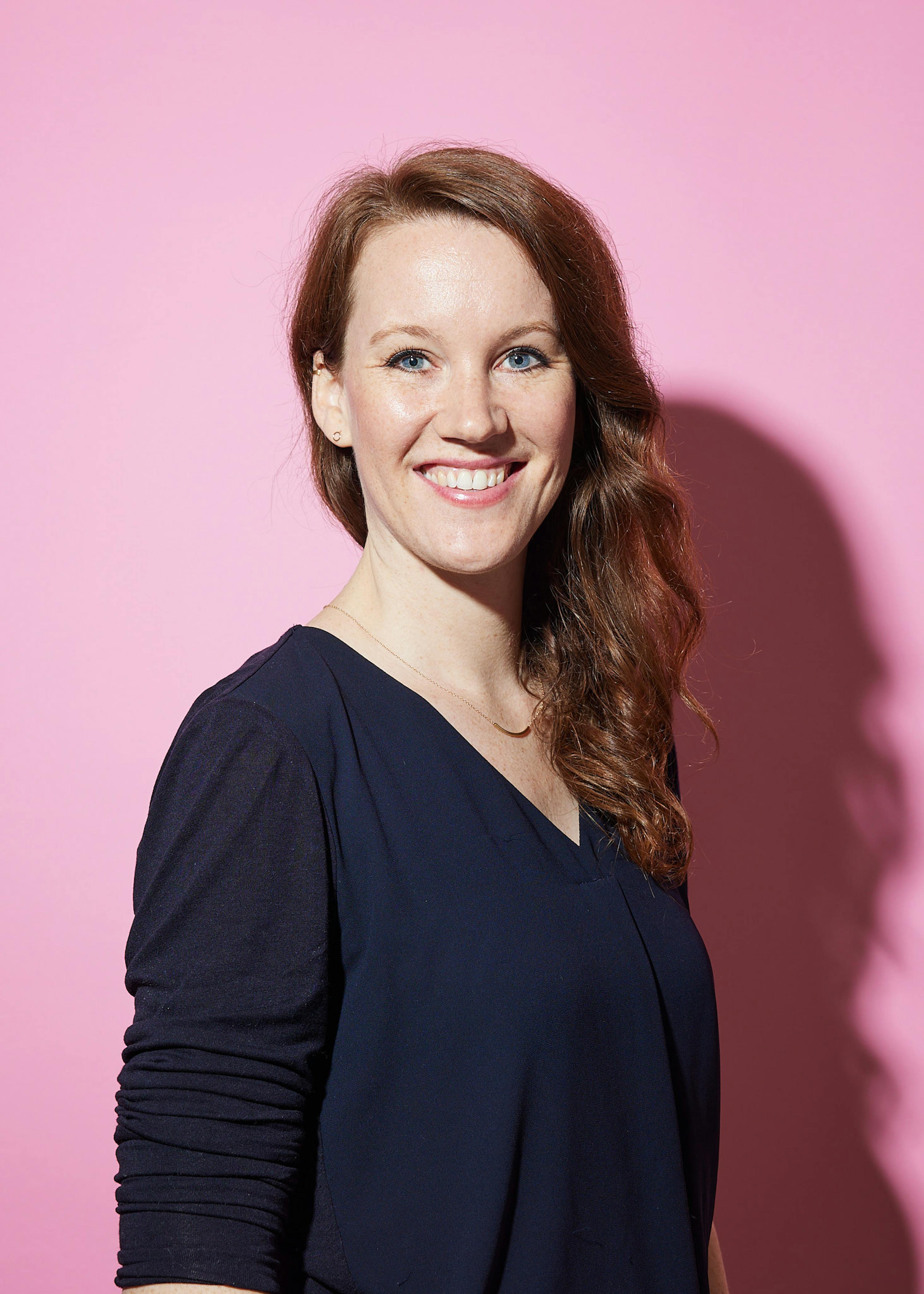 Eine Frau lächelt vor einem rosa Hintergrund und trägt ein blaues Hemd