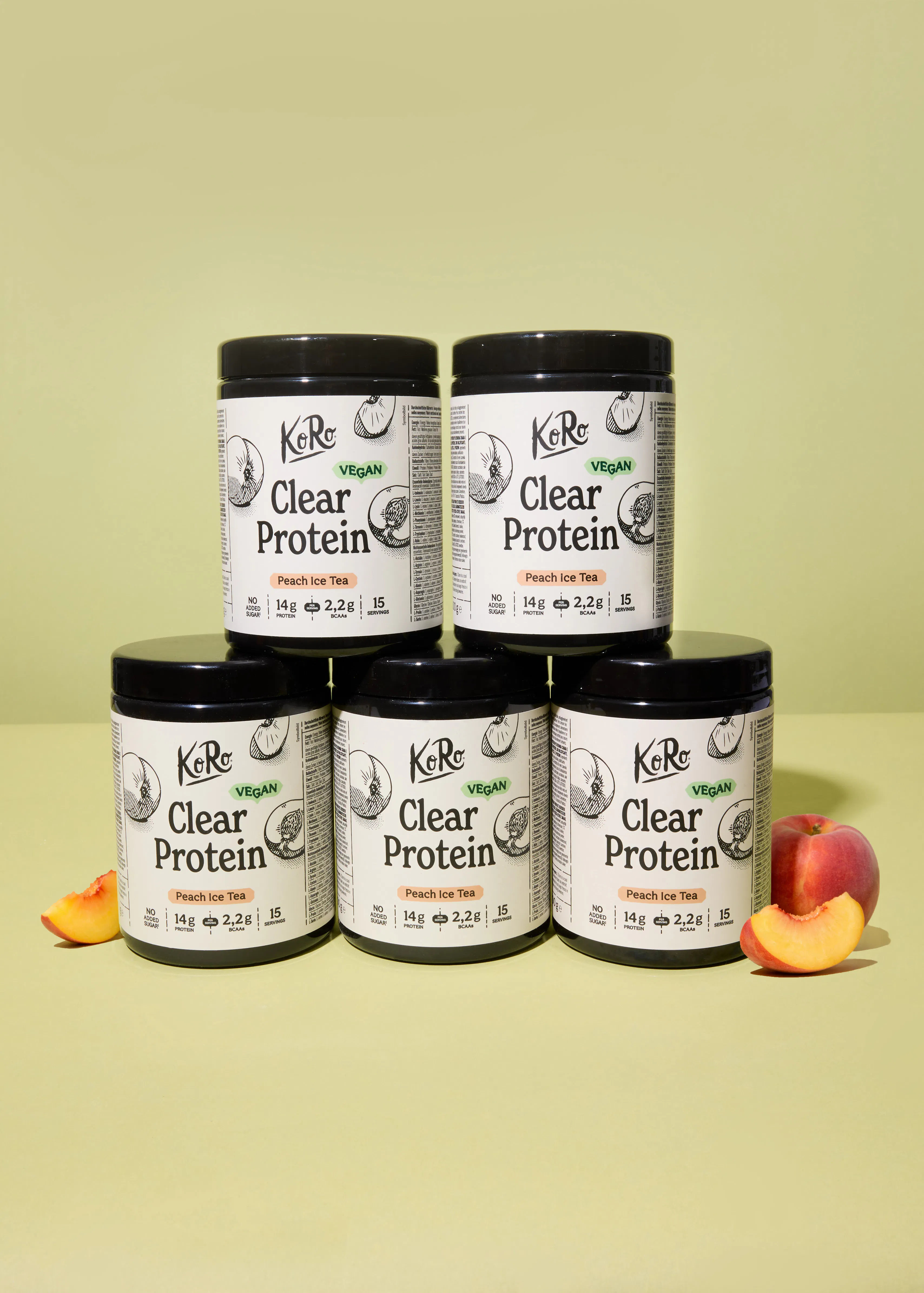 five black jars of koro vegan clear protein peach ice tea are stacked on a light green background, accompanied by fresh peaches and peach slices.