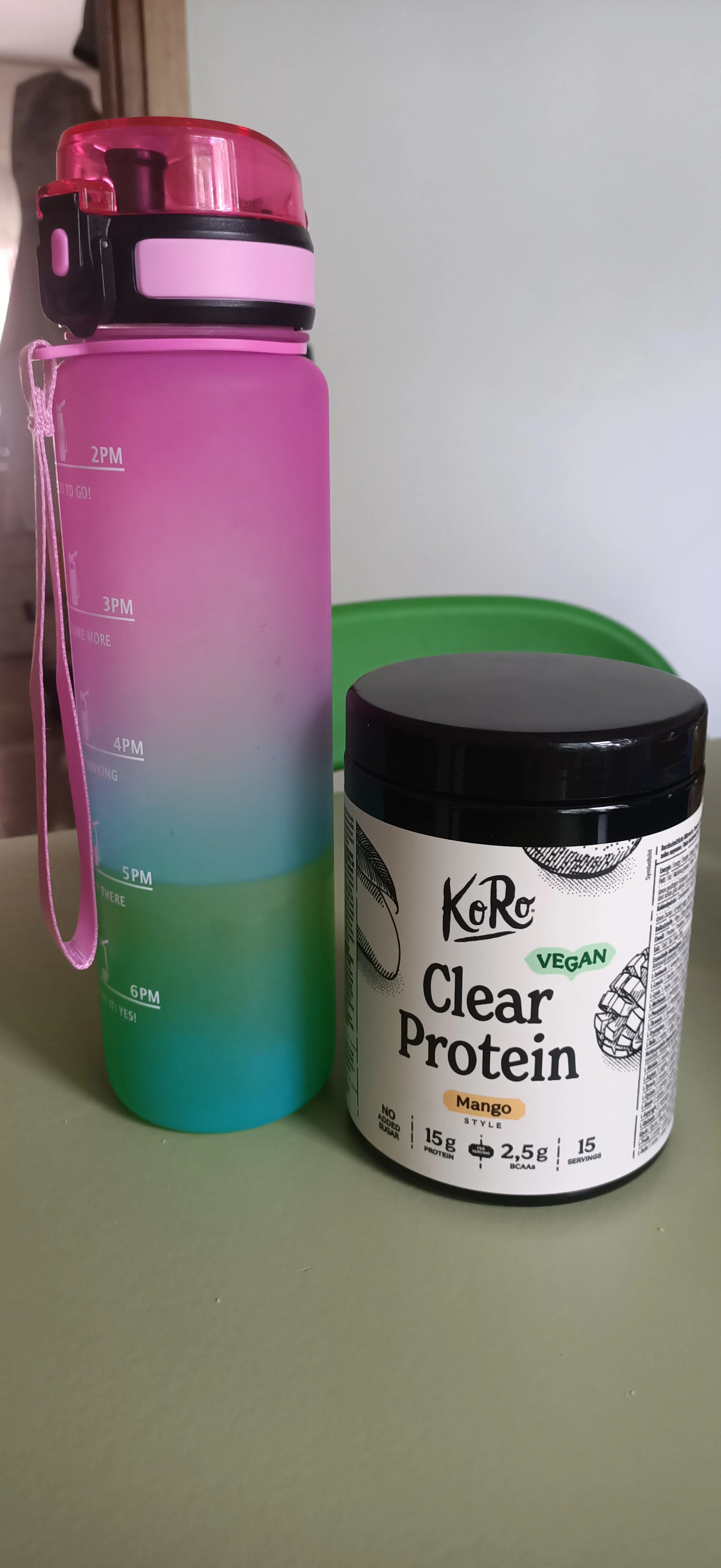 eine pink-grüne wasserflasche mit zeitmarkierungen steht neben einem behälter mit koro clear protein mangopulver.