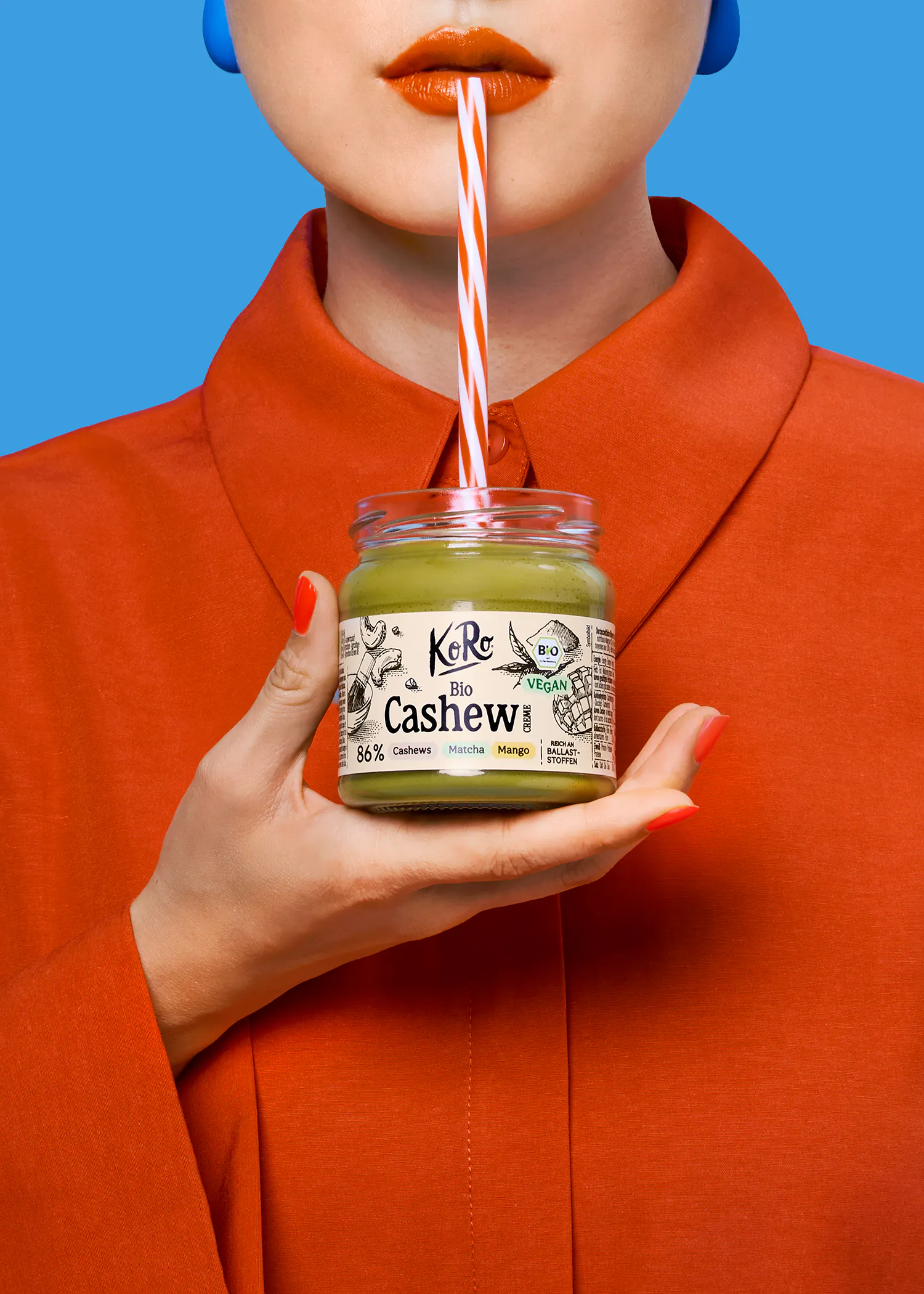 eine person in einem orangefarbenen hemd mit orangefarbenem lippenstift trinkt mit einem gestreiften strohhalm aus einem glas koro bio cashew creme vor blauem hintergrund.