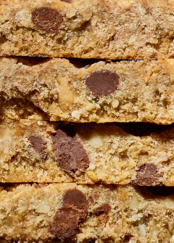 gros plan sur une pile verticale de quatre barres de biscuits dorées, parsemées de pépites de chocolat et de morceaux de noix.