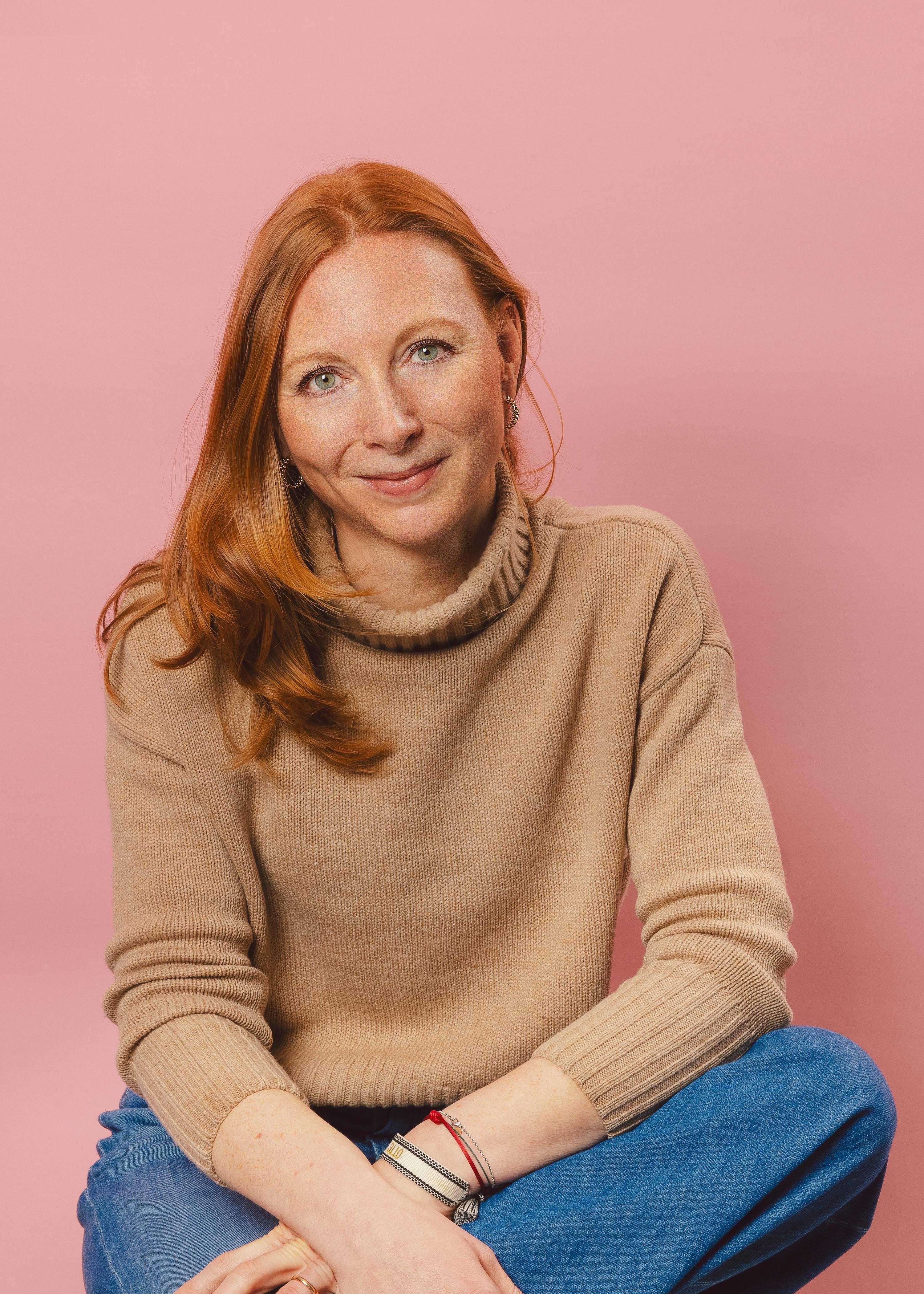a red-haired woman in a beige turtleneck sweater and blue jeans smiles at the camera against a pink background.