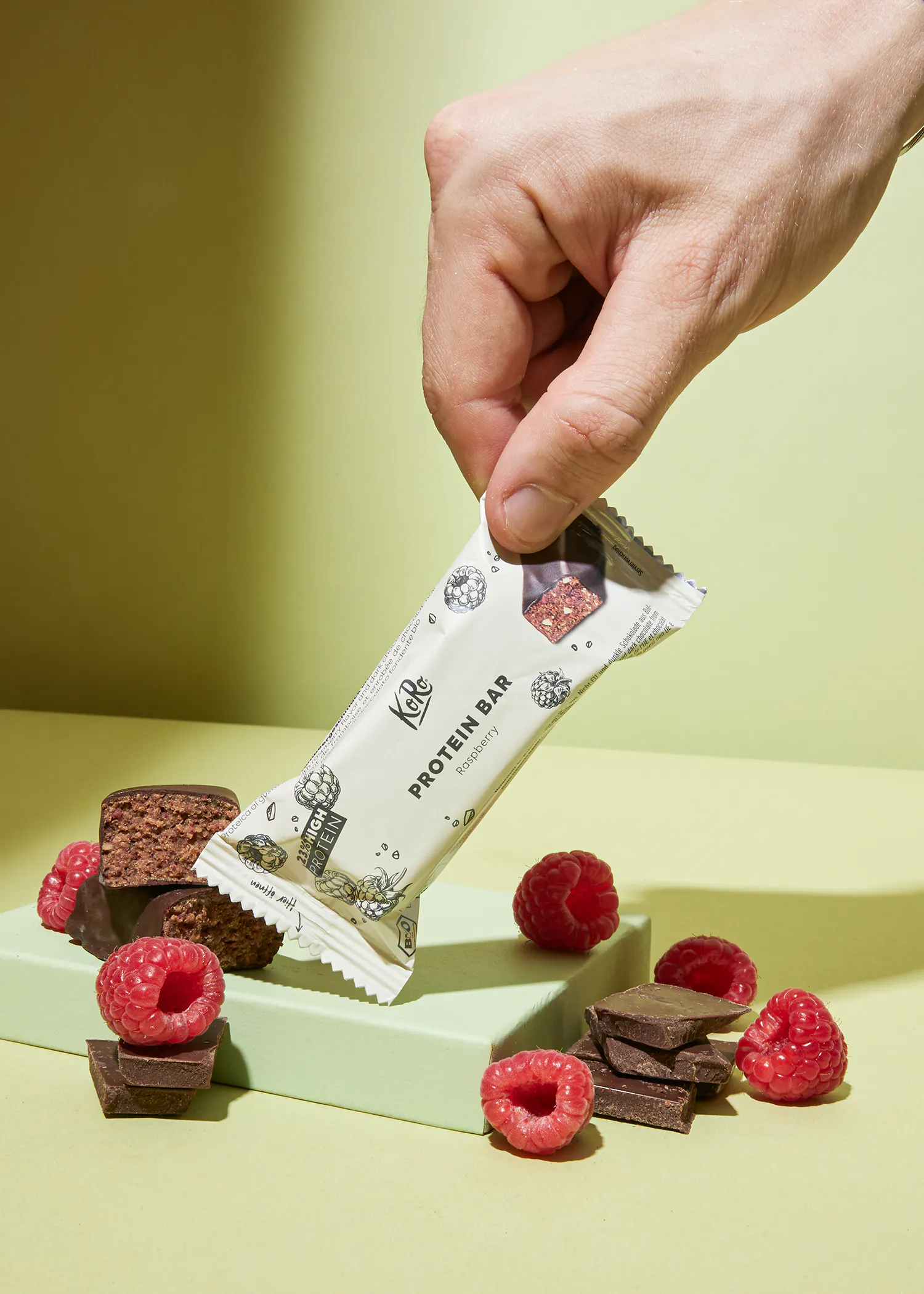 una persona sostiene una barra de proteína frente a frambuesas y chocolate