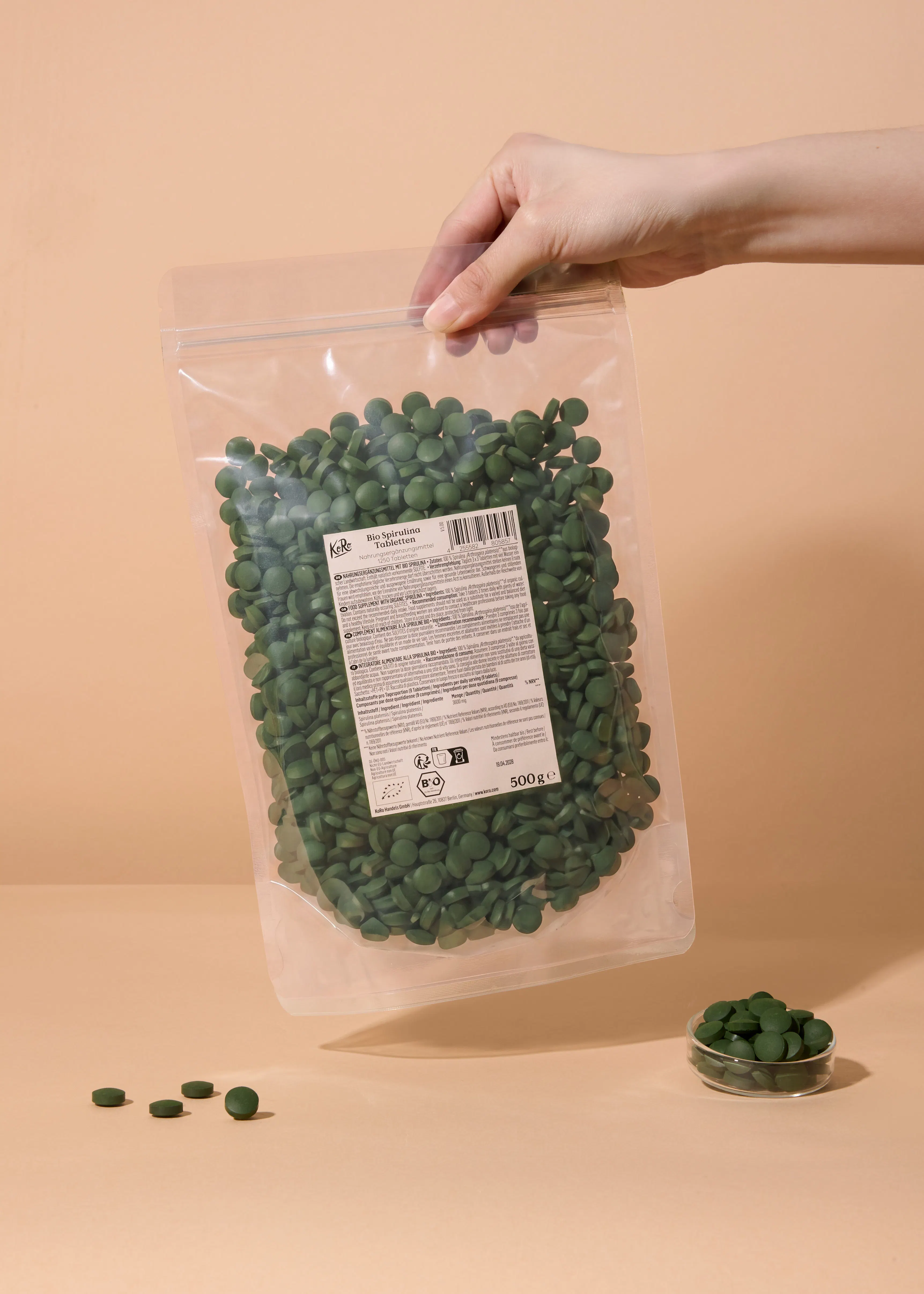 eine hand hält einen durchsichtigen beutel mit grünen bio spirulina tabletten, von denen einige auf einer beigen oberfläche und in einer kleinen glasschale liegen.